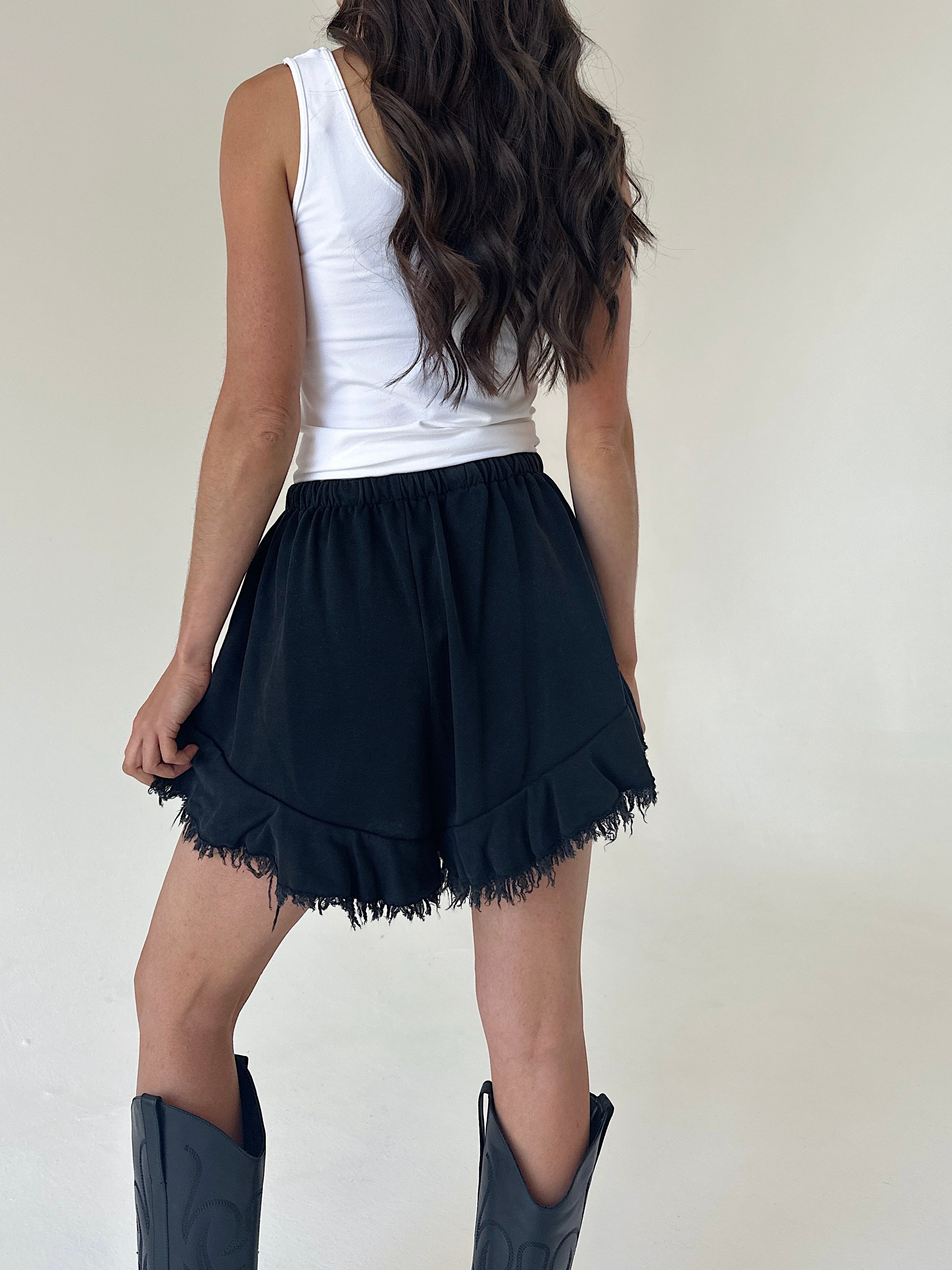 A woman shown from the back wearing a white sleeveless top, black ruffled shorts, and dark cowboy boots against a plain light background.
