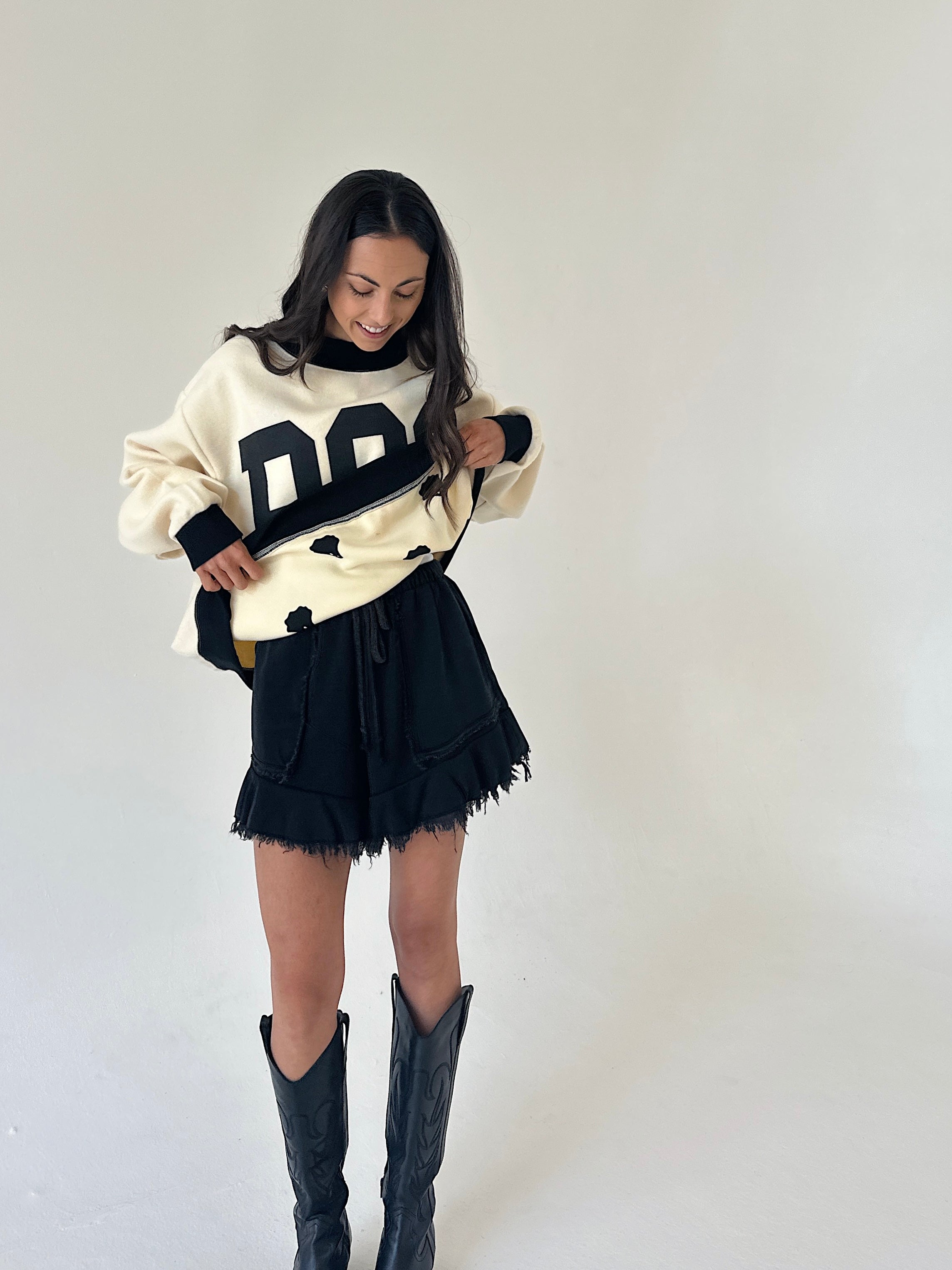 A woman modeling a cream and black knit sweater with large letter graphics, paired with black fringed shorts and black cowboy boots, against a plain off-white studio background.