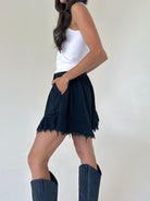 A woman wearing a black frayed terry shorts with a white tank top and dark cowboy boots, shown from a side-back angle against a plain off-white background.
