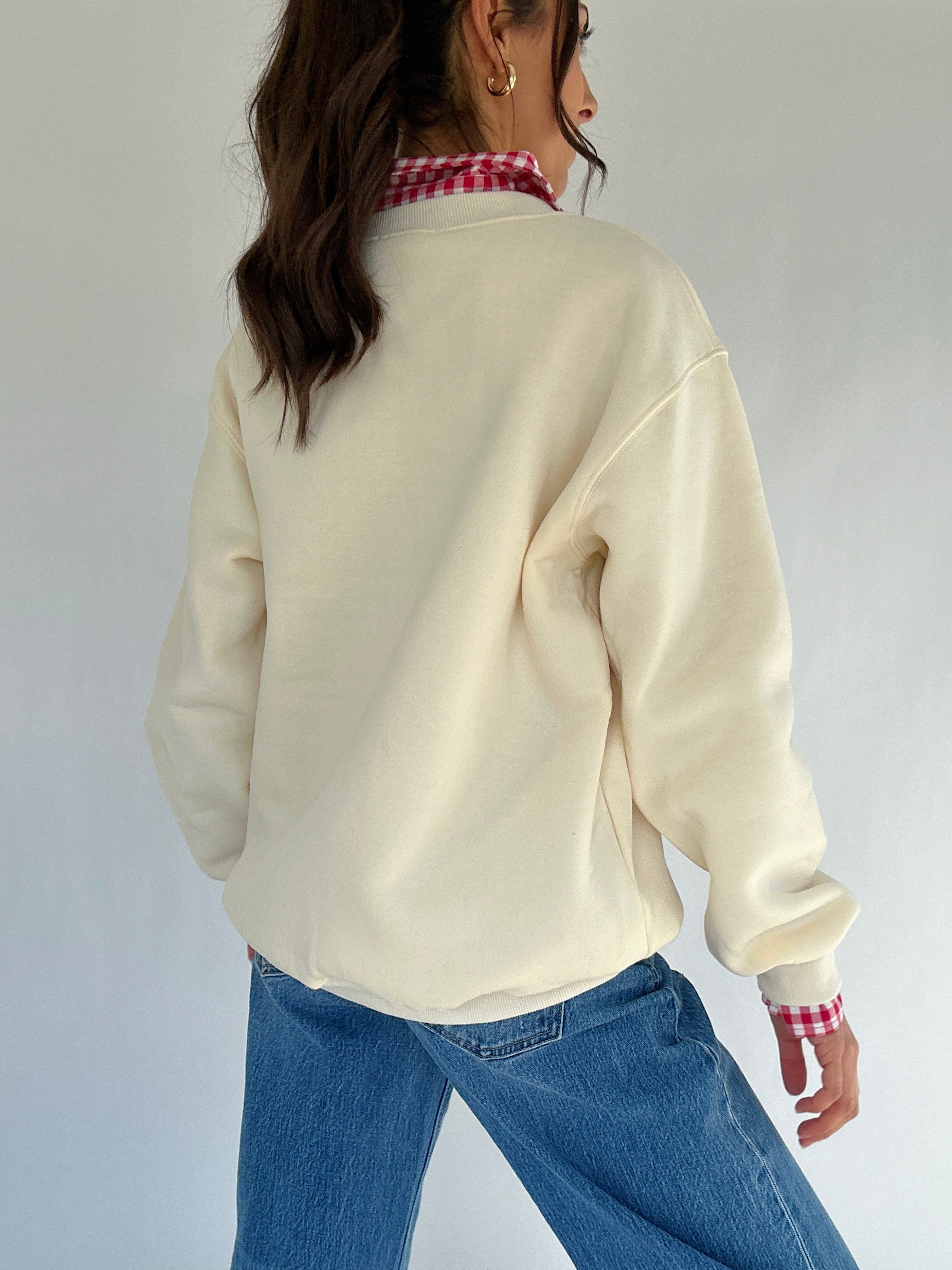 Person wearing a cream sweatshirt with a checkered collar and blue jeans on a plain background