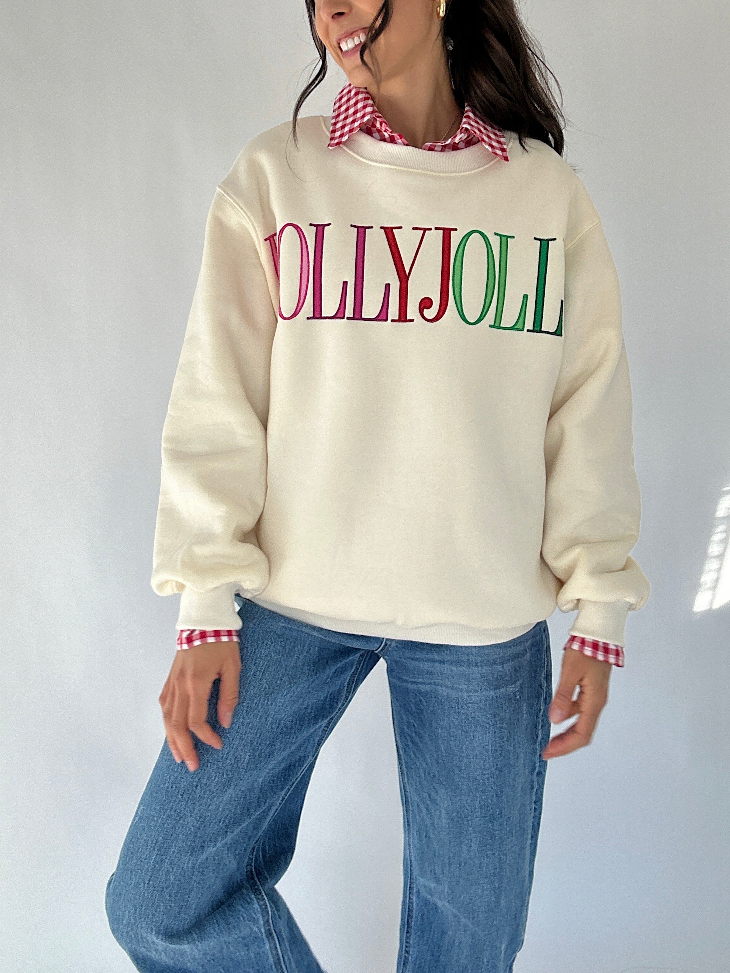 Person wearing a cream sweatshirt with 'HOLLY JOLLY' text, blue jeans, and a checkered shirt on a white background