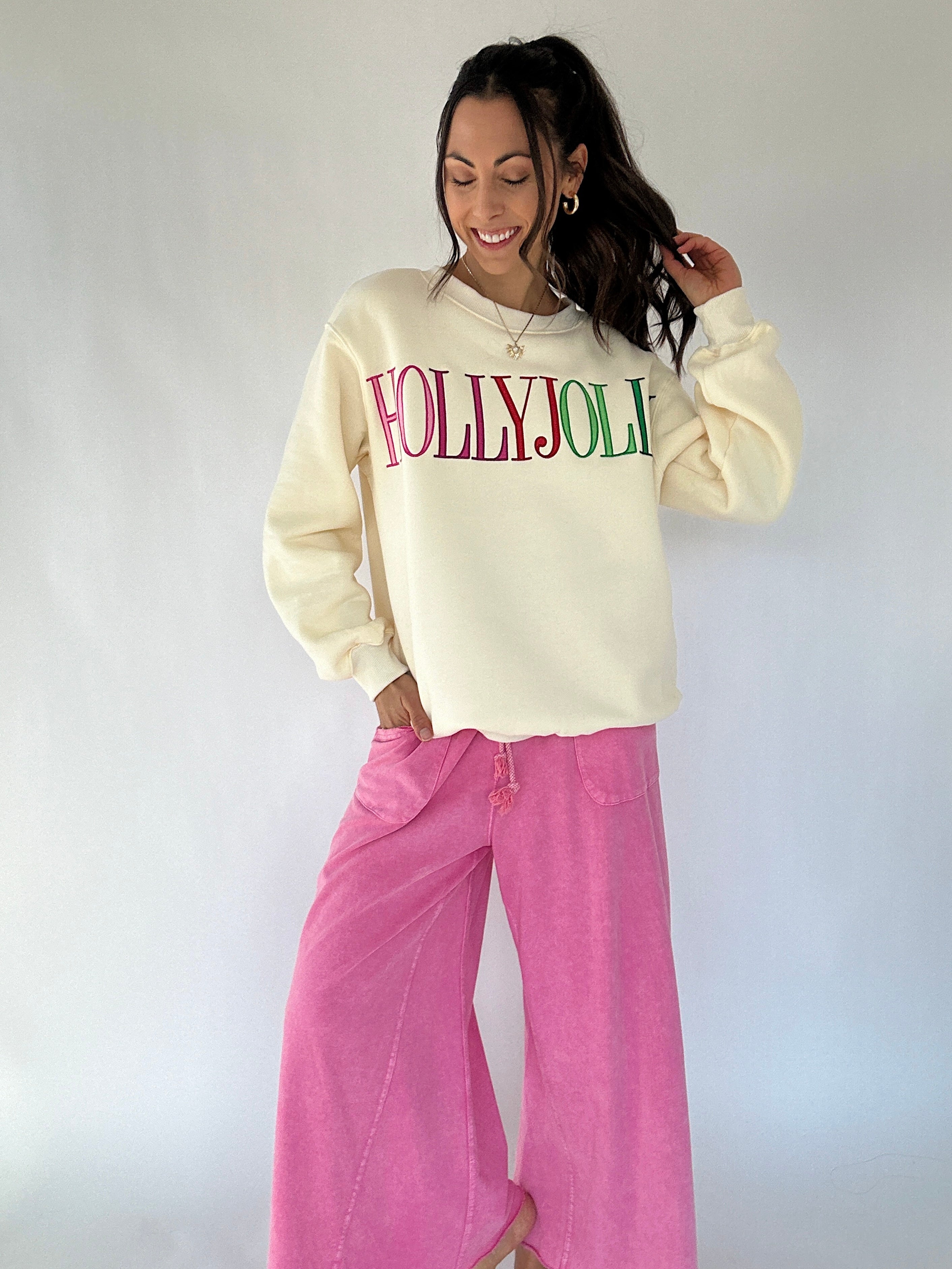 Woman wearing a cream sweatshirt with 'HOLLYJOLLY' text and pink pants against a white background