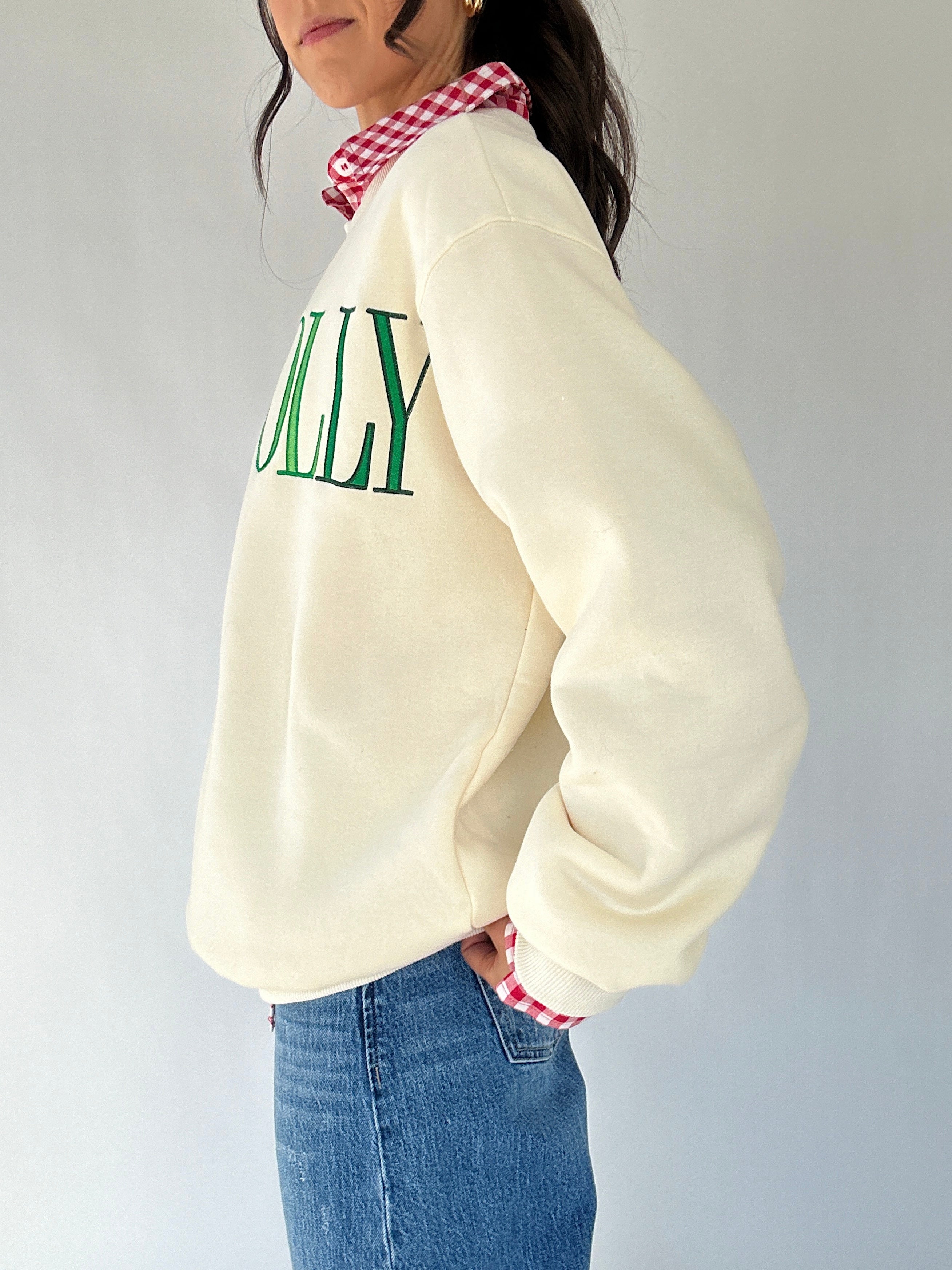 Person wearing a cream sweatshirt with green text, blue jeans, and a checkered under shirt on a plain background