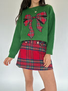 Woman wearing a green sweater with a red plaid bow design and matching red plaid skort on a white background