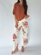 A woman modeling a rust-colored knit poncho sweater and cream wide-leg pants with a red and gray floral pattern, standing against a light gray studio background.