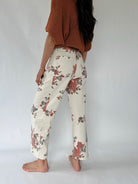 A person shown from the back wearing cream wide-leg pants with a muted floral print in red, brown, and gray, paired with a rust-colored knit top, standing barefoot against a light gray studio background.
