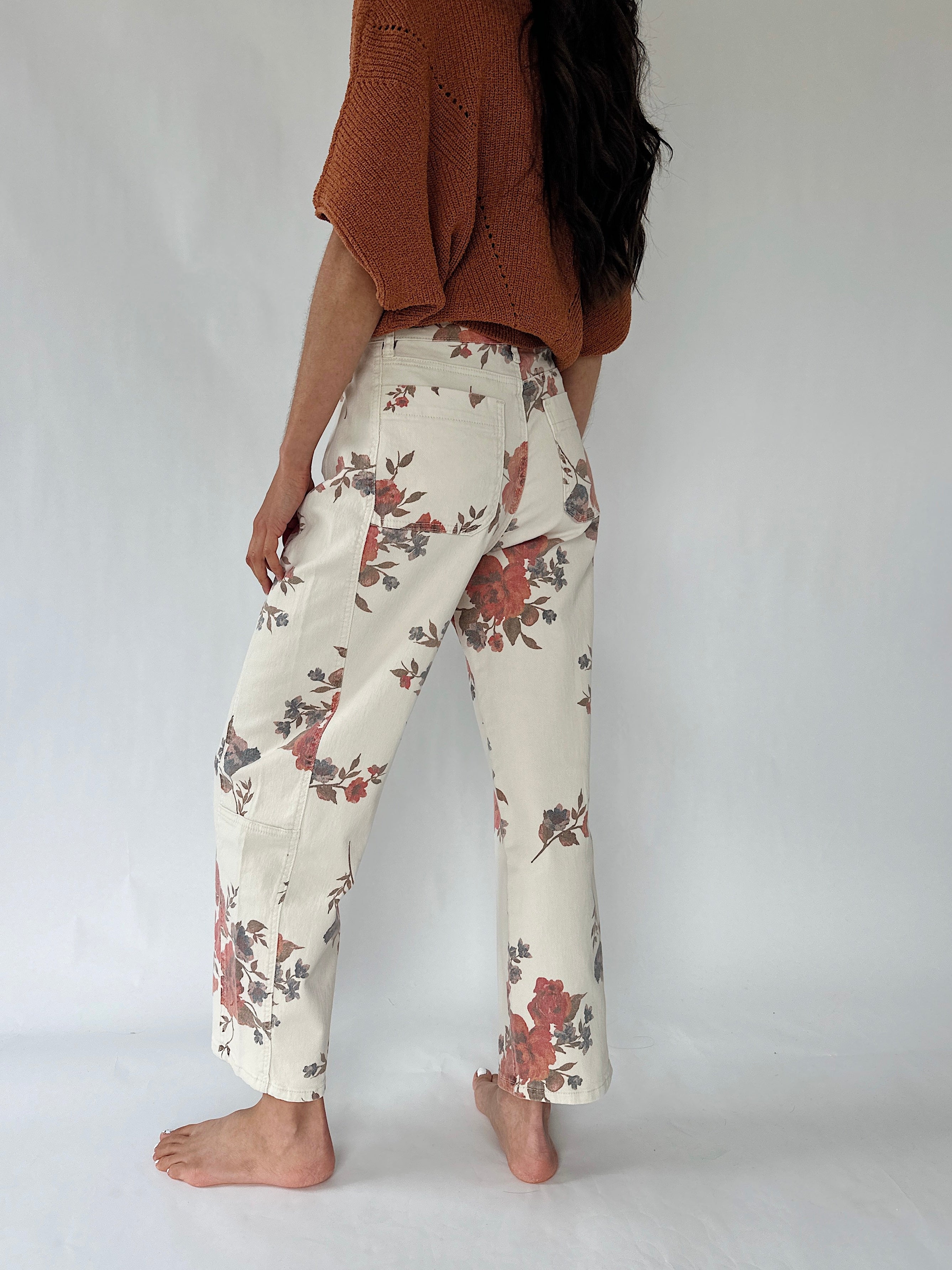 A person shown from the back wearing cream wide-leg pants with a muted floral print in red, brown, and gray, paired with a rust-colored knit top, standing barefoot against a light gray studio background.