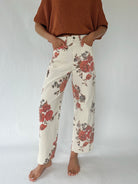 A person wearing cream wide-leg pants with a red and brown floral print, paired with a rust-colored knit top, standing against a light gray studio background.