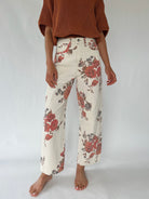 A model is shown from the chest down wearing high-waisted, wide-leg cropped pants with a red and brown floral print on an off-white base, paired with a rust-colored knit sweater.