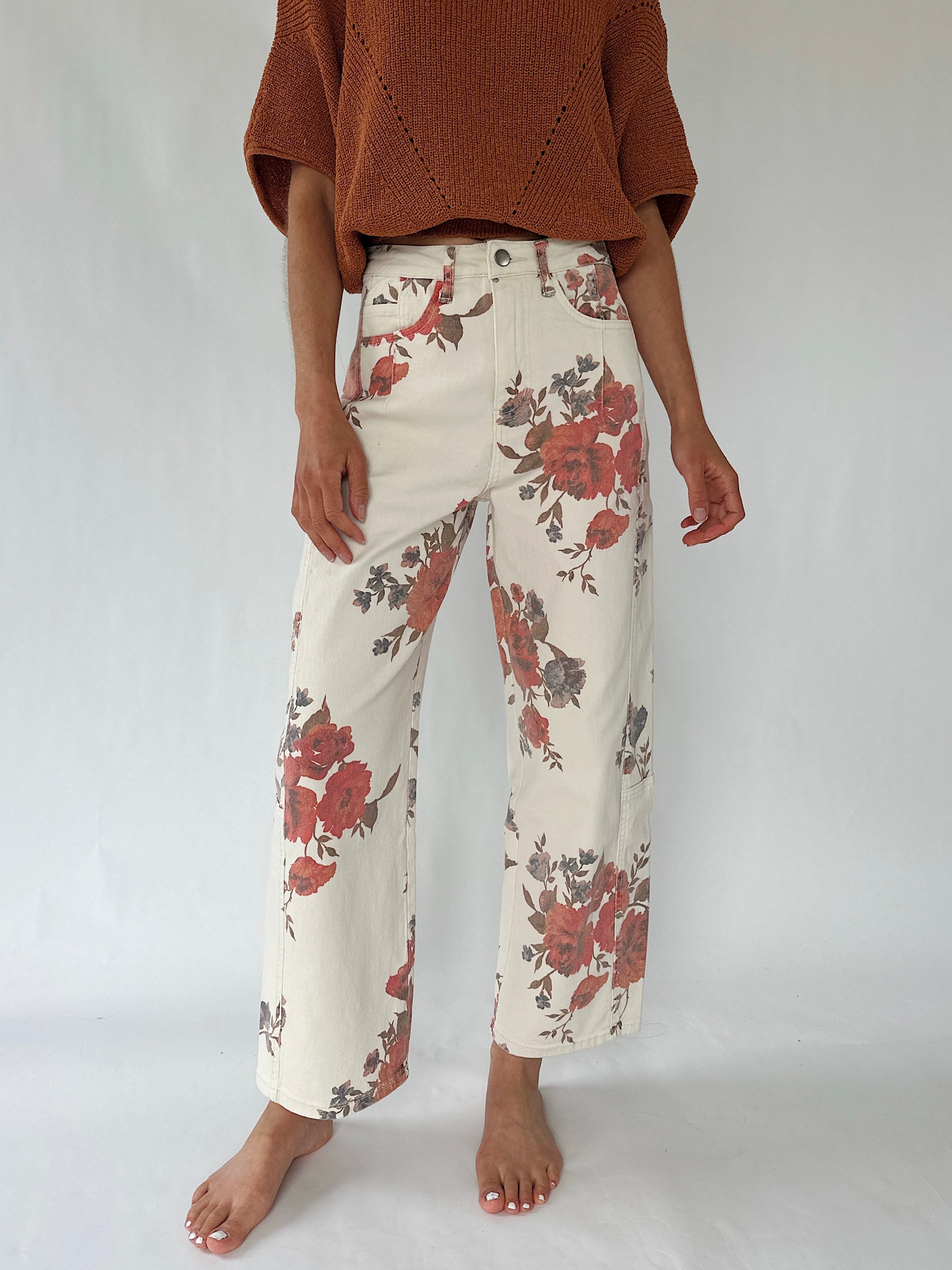 A model is shown from the chest down wearing high-waisted, wide-leg cropped pants with a red and brown floral print on an off-white base, paired with a rust-colored knit sweater.