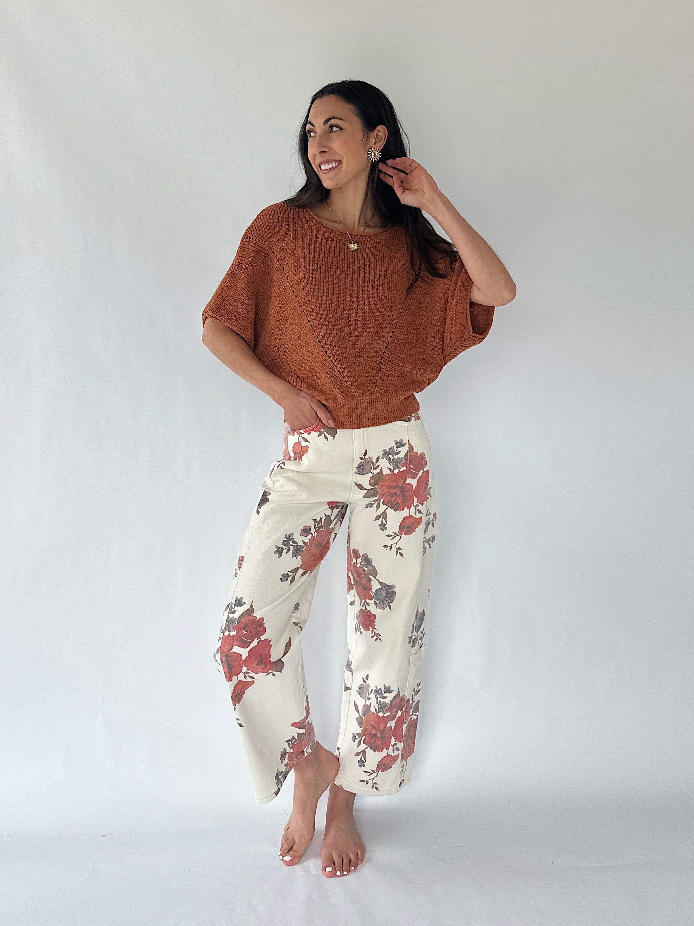 A woman modeling a rust-colored knit poncho top and cream wide-leg pants with a red and gray floral pattern, standing against a white studio backdrop.