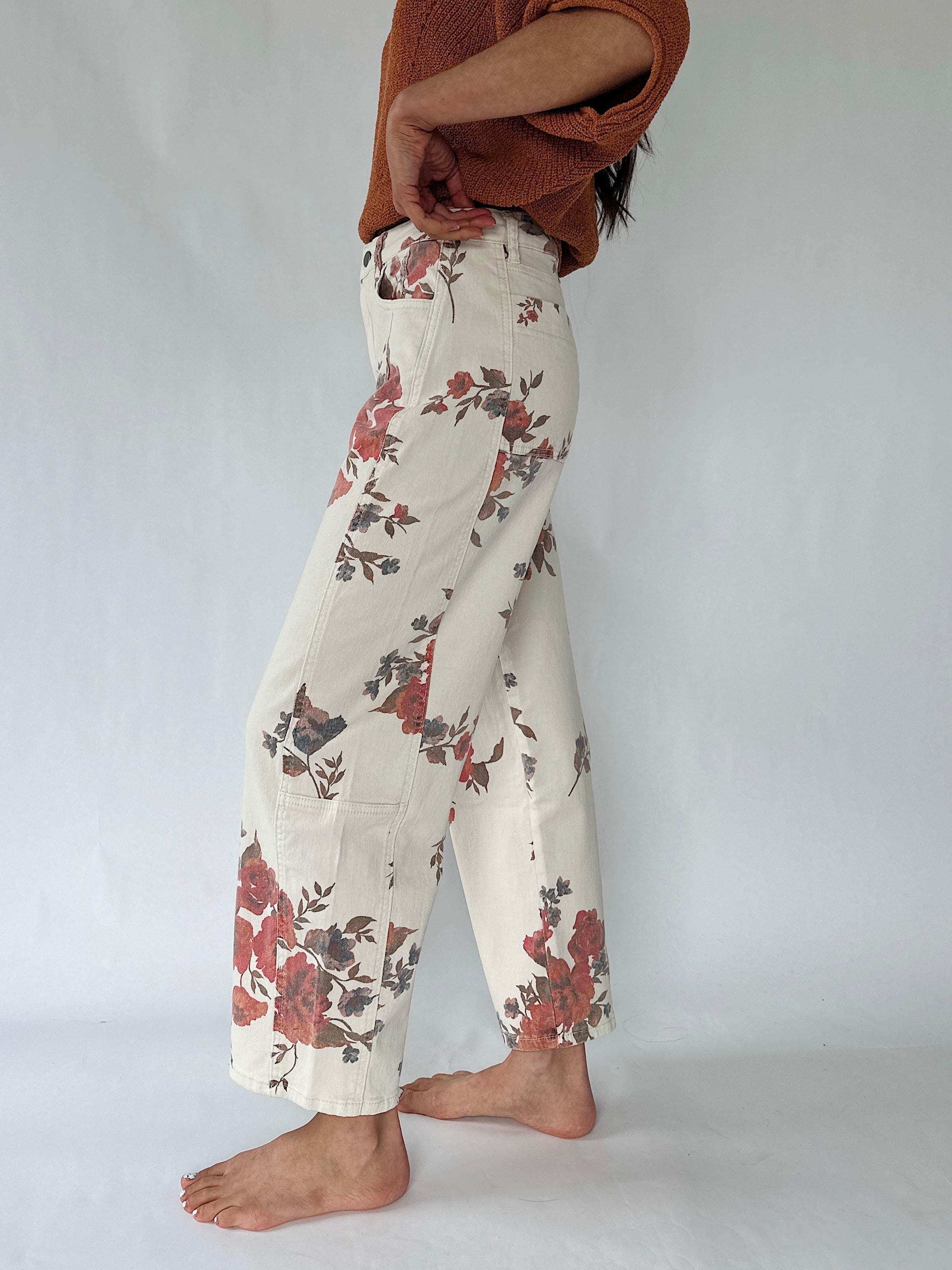 A person shown from the waist down wearing cream wide-leg pants with a red and brown floral print, paired with a rust-colored knit top, standing barefoot against a white studio background.