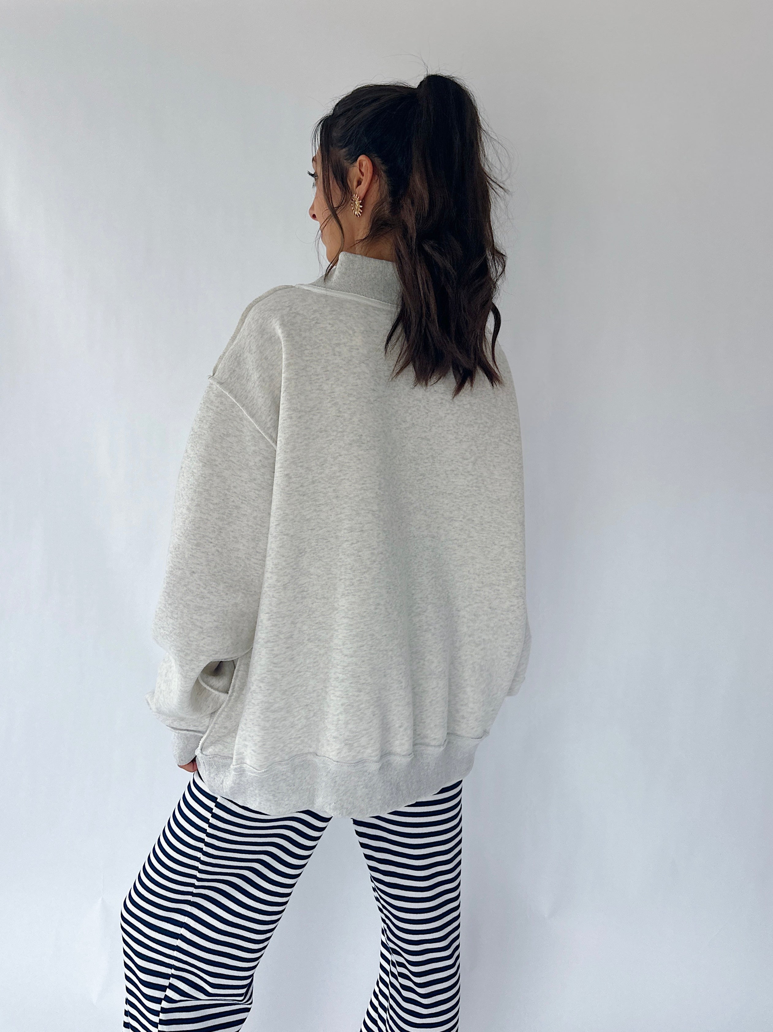A woman shown from the back wearing an oversized light gray heather sweatshirt and black-and-white striped pants, standing against a plain light gray wall.