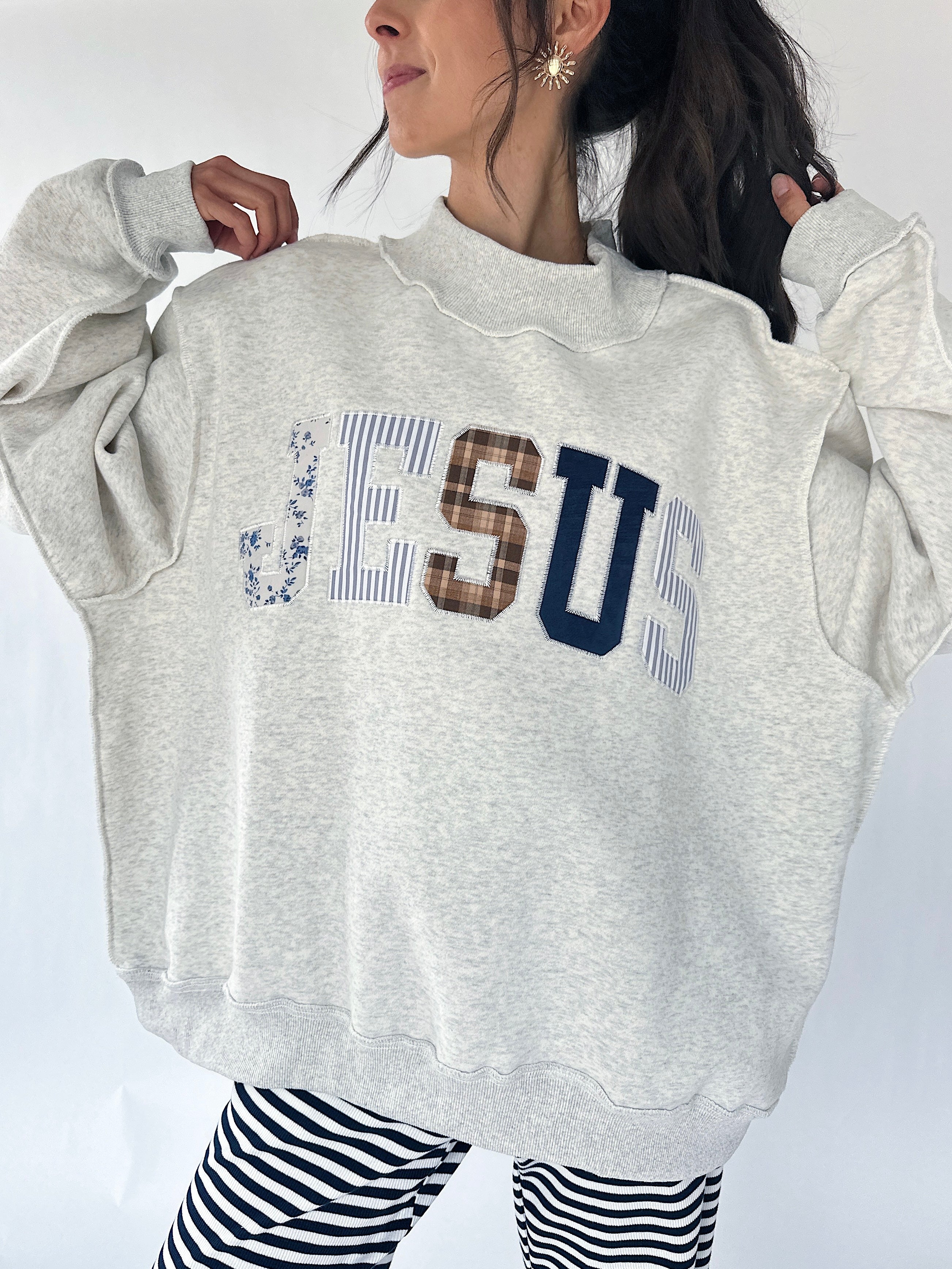 A woman wearing an oversized light gray sweatshirt with 'JESUS' printed across the chest in mixed patterned lettering, paired with black and white striped pants.