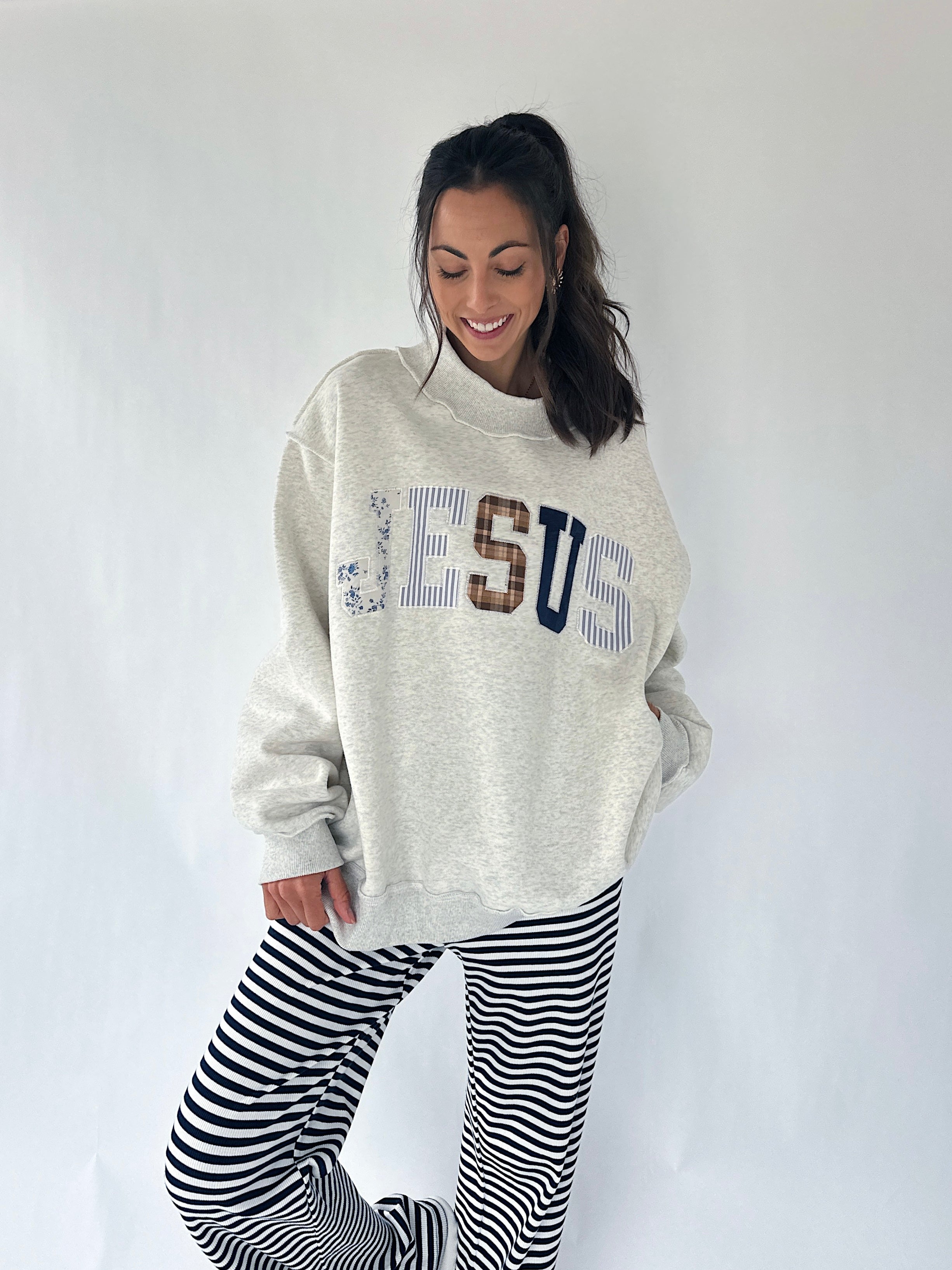 A woman modeling an oversized cream heather sweatshirt with 'JESUS' written across the front in blue, brown, and navy lettering, paired with black and white horizontally striped pants, against a plain white studio background.