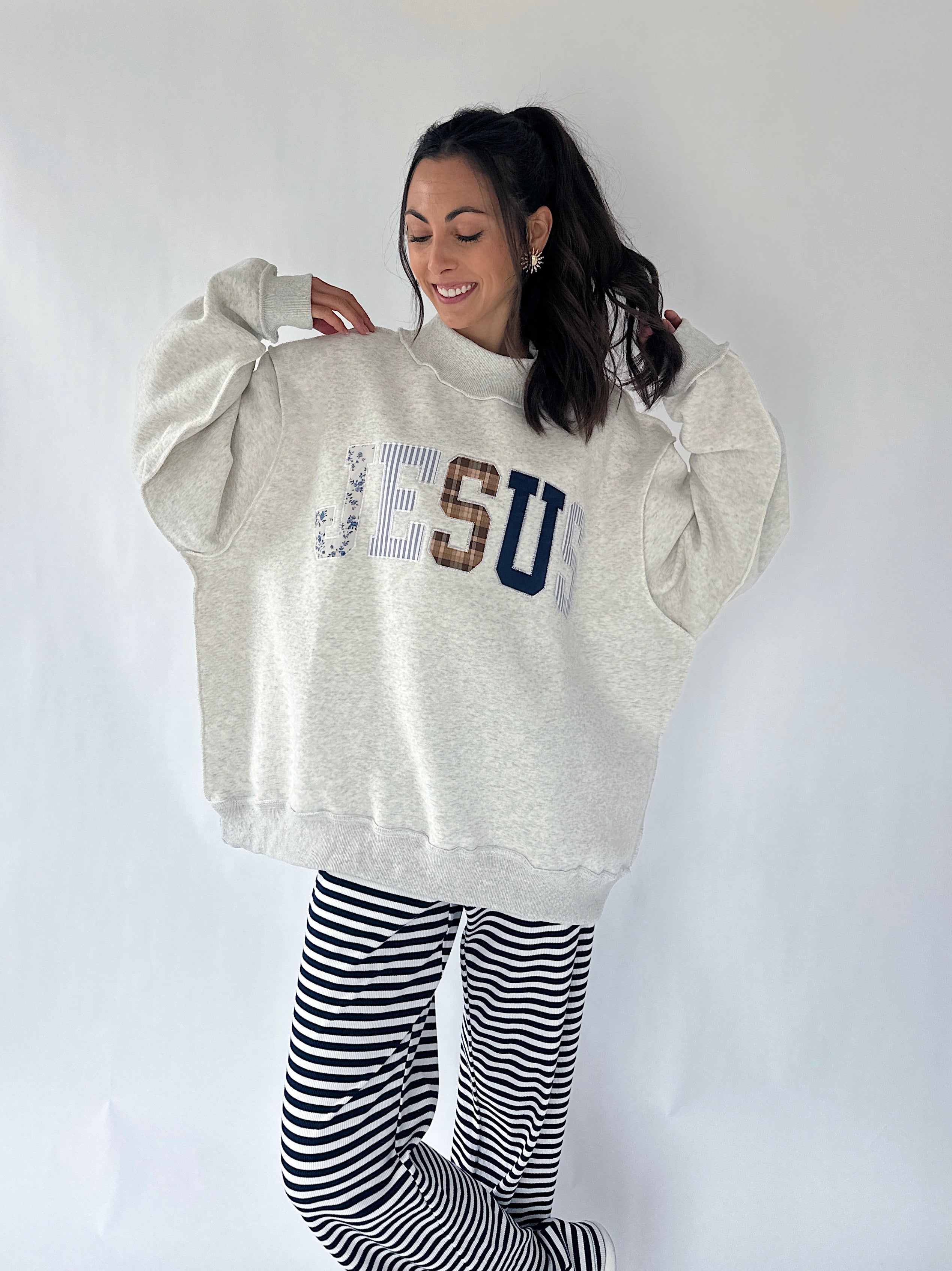 A woman modeling an oversized light heather gray sweatshirt with 'JESUS' printed across the chest in mixed textures, paired with black and white striped pants, against a plain white studio background.