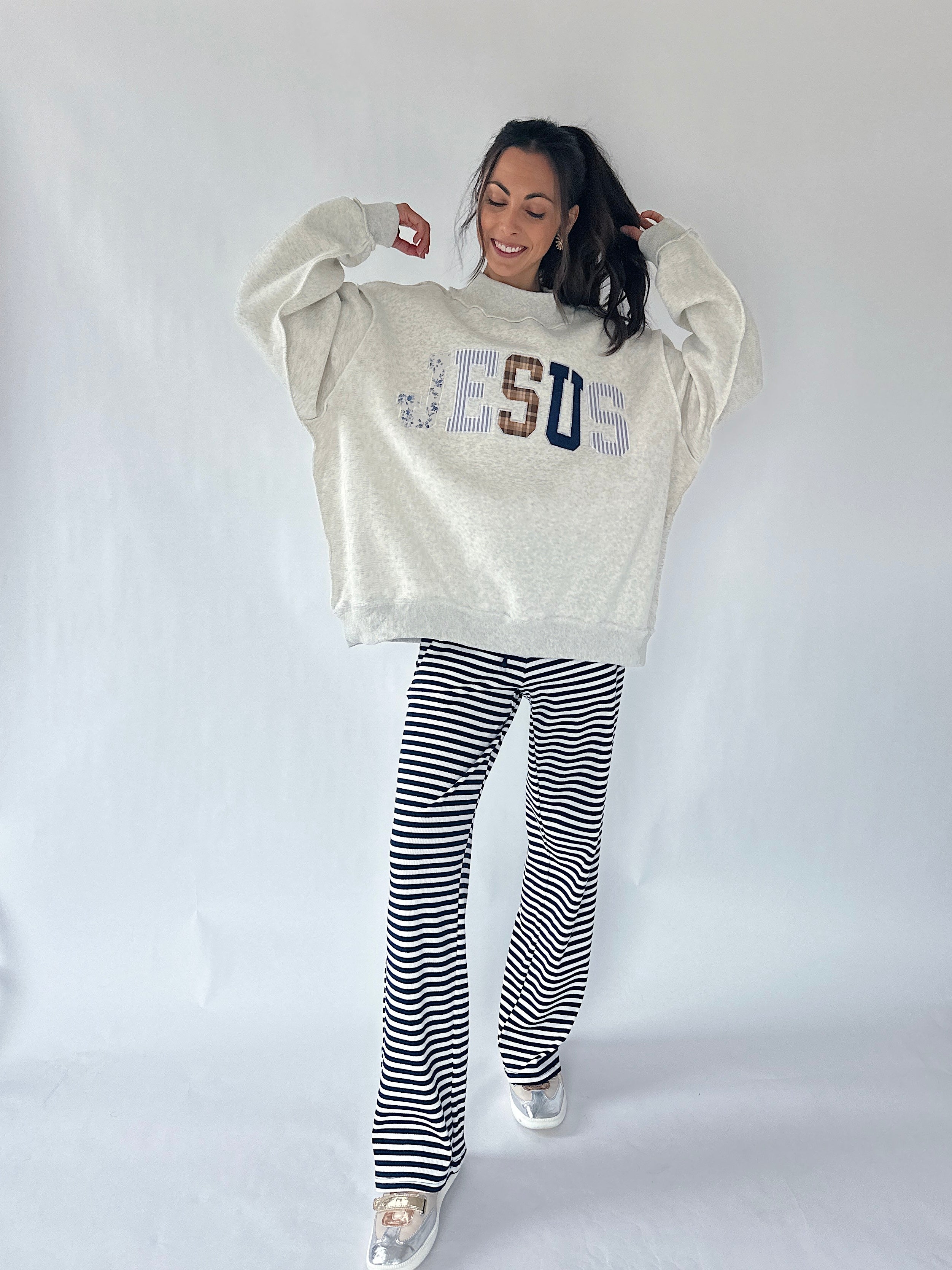 A woman modeling a cream-colored oversized sweatshirt with 'JESUS' written across the chest in blue, brown, and white lettering, paired with black and white horizontally striped pants, against a plain white studio background.