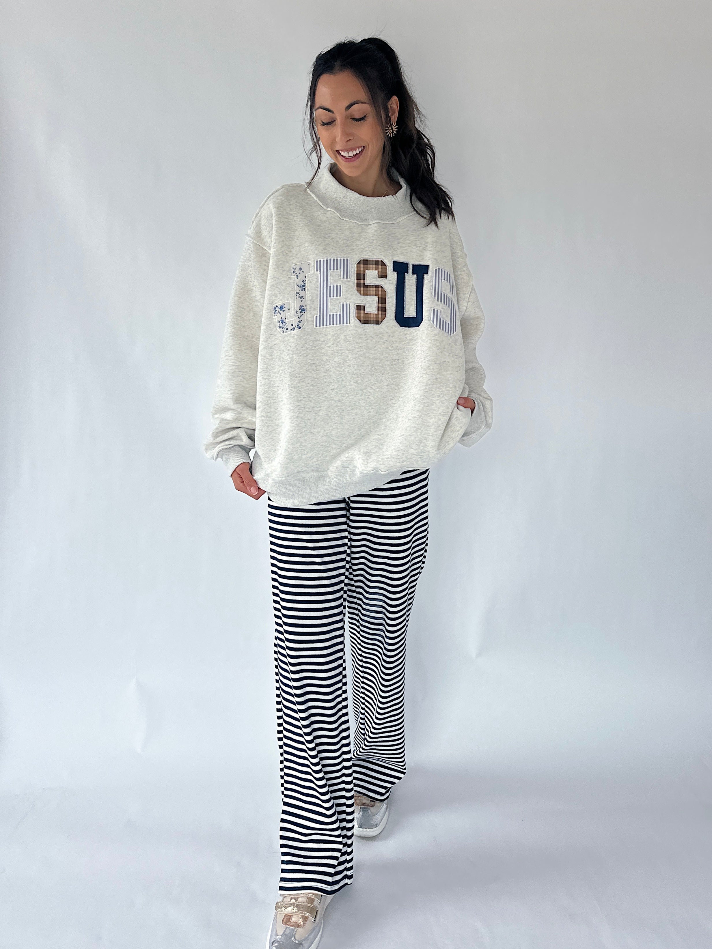 A woman modeling a cream-colored sweatshirt with 'JESUS' spelled out in decorative letters, paired with black and white striped wide-leg pants, against a plain light gray studio background.