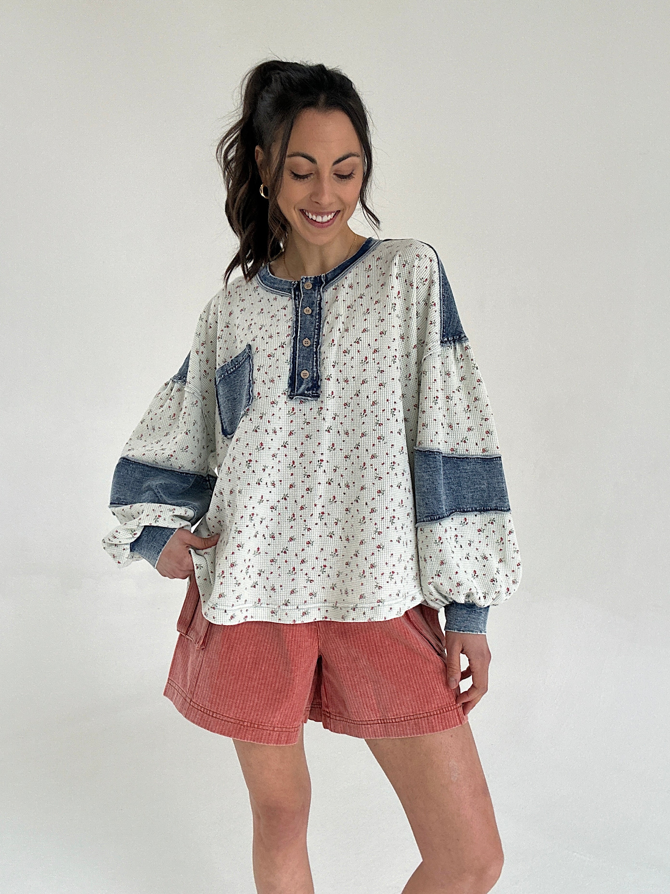 Woman wearing a patterned long sleeve top with denim patches and rust shorts on a white background