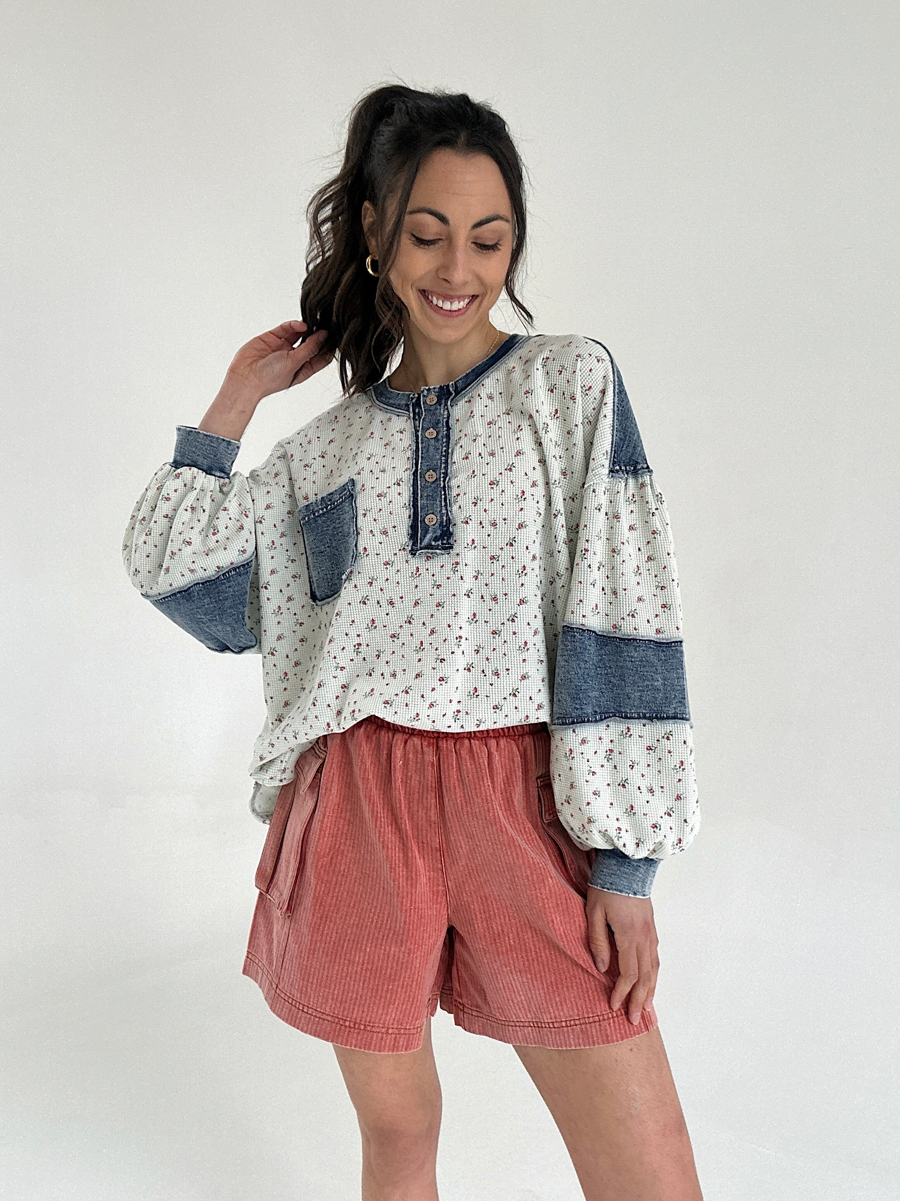 Woman wearing a floral long sleeve top with denim accents tucked into rust red shorts on a white background