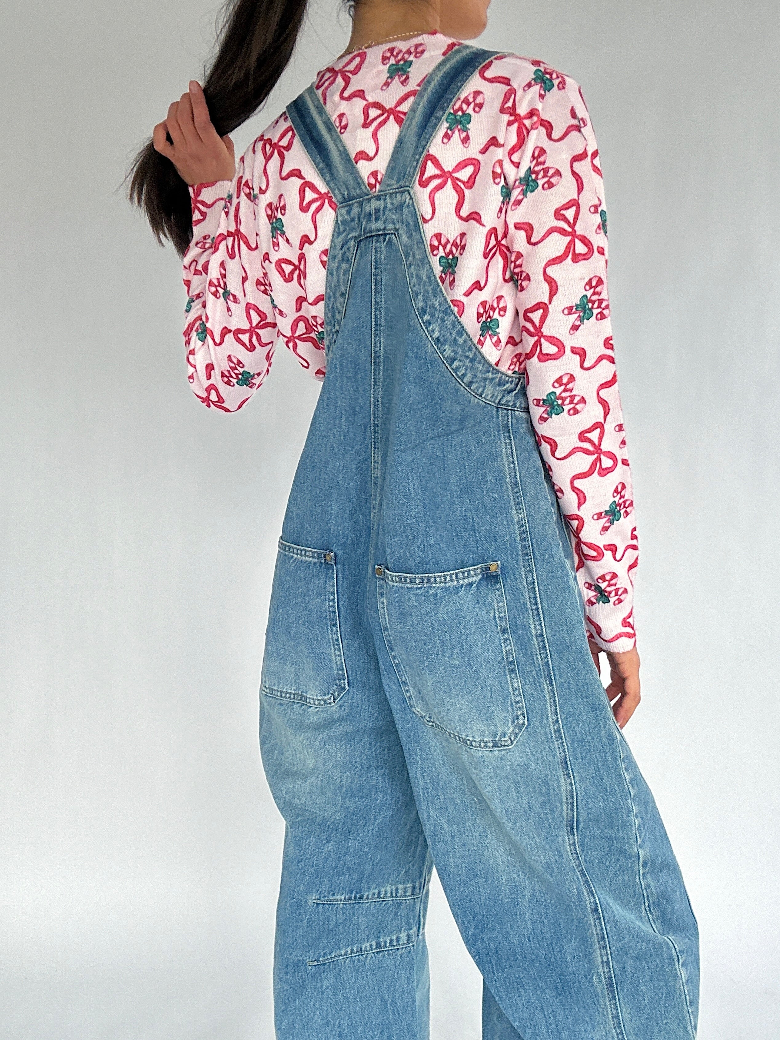 Person wearing a pink candy cane shirt and blue denim overalls on a plain background showing back