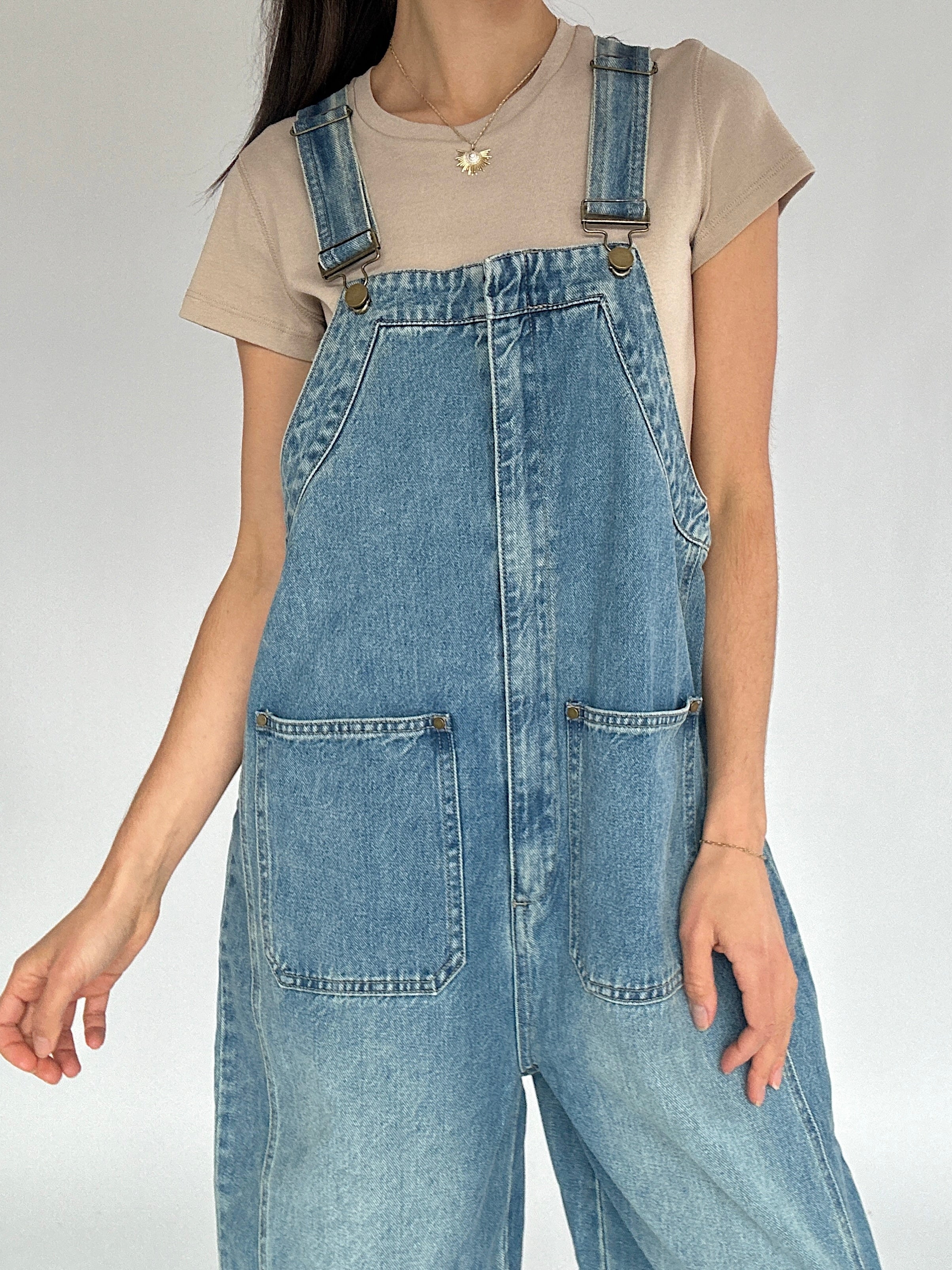 Person wearing blue denim overalls on a plain background showing buckle strap and front pocket details