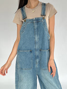 Person wearing blue denim overalls on a plain background showing buckle strap and front pocket details