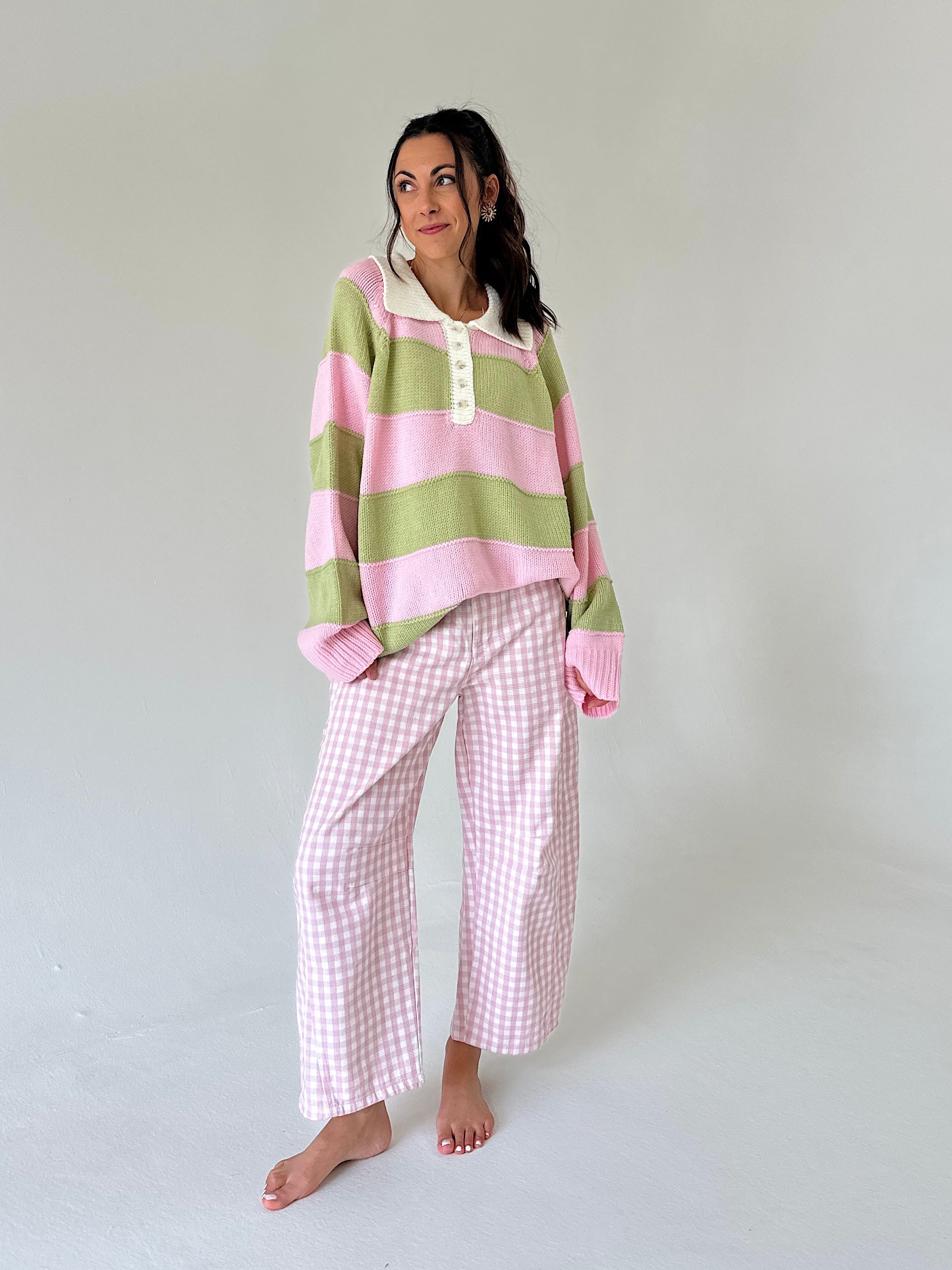 A woman modeling a cozy outfit consisting of a pink and light green striped long-sleeve henley sweater and pink-and-white gingham wide-leg pants, set against a plain light gray studio background.