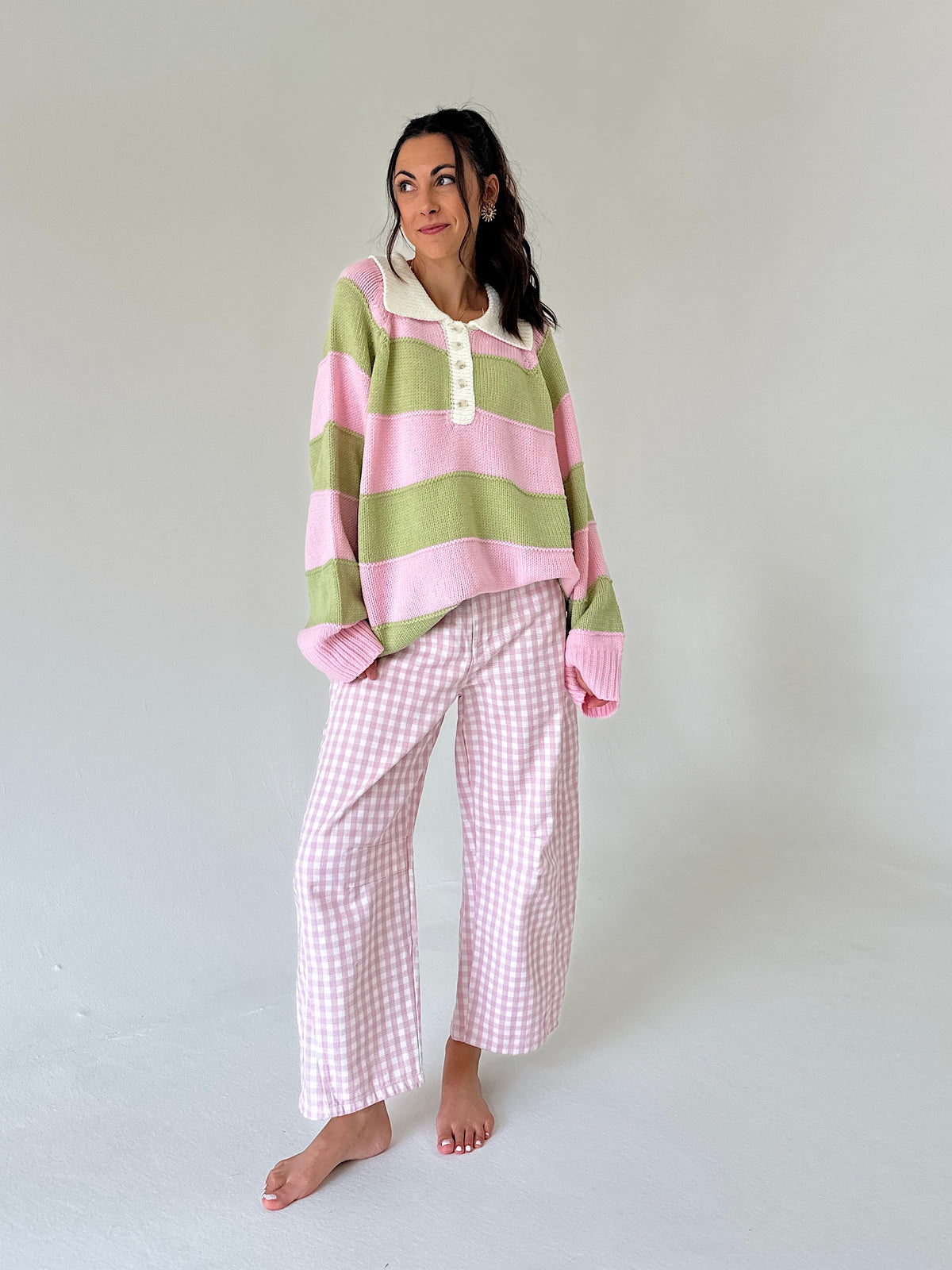 A woman modeling a cozy outfit consisting of a pink and light green striped long-sleeve henley sweater and pink-and-white gingham wide-leg pants, set against a plain light gray studio background.