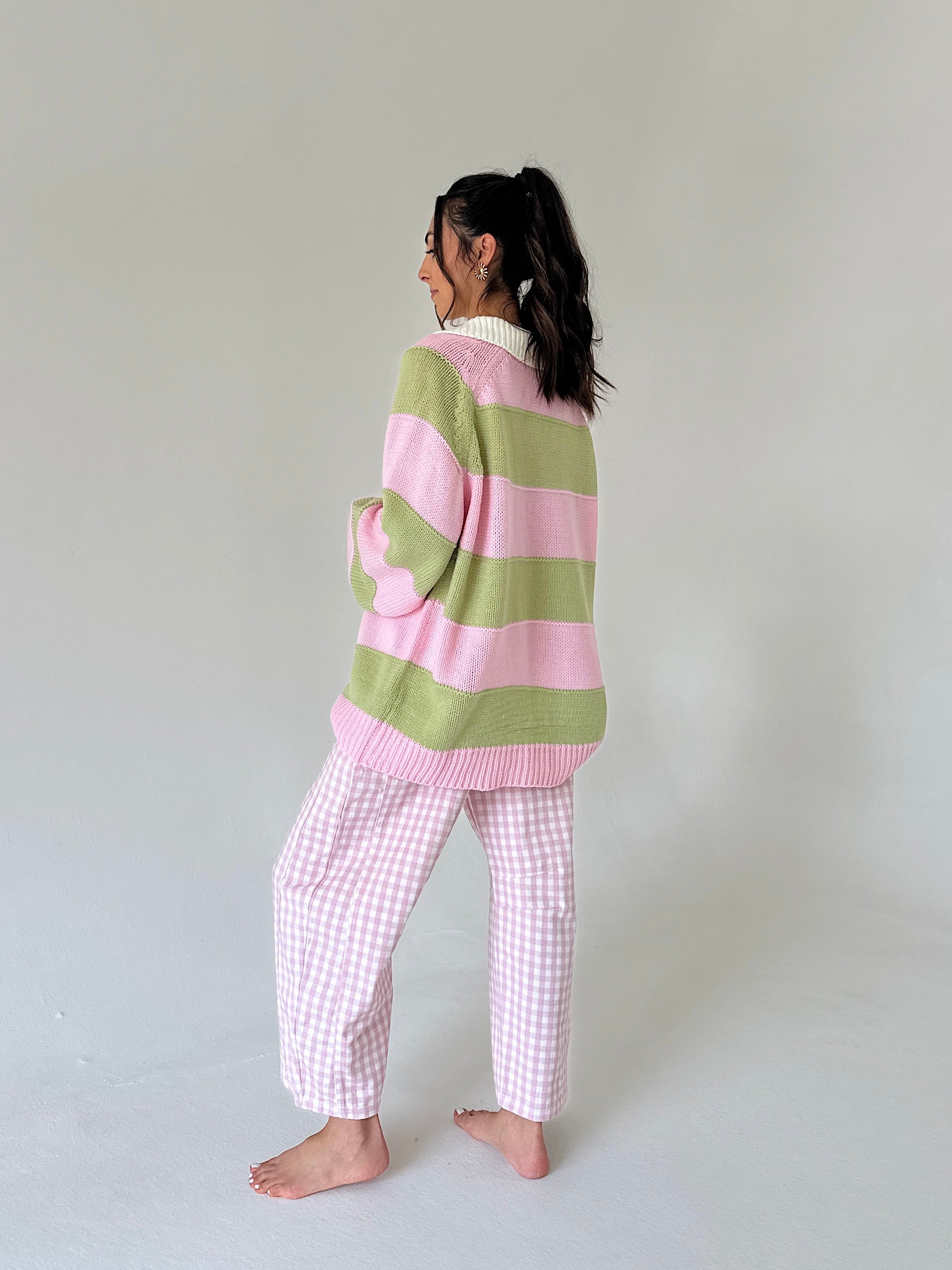 A woman stands barefoot in profile wearing a pastel striped knit sweater with pink and light green horizontal bands, paired with pink-and-white gingham pajama pants, against a plain light gray studio background.