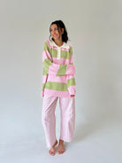 A woman stands barefoot against a plain light gray studio background wearing a pink and sage green striped long-sleeve henley top with a white collar and matching pink-and-white gingham pants.