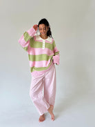 A woman modeling a pink and light green striped long-sleeve polo sweater with a white collar, paired with pink and white gingham check pants, against a plain light gray studio background.