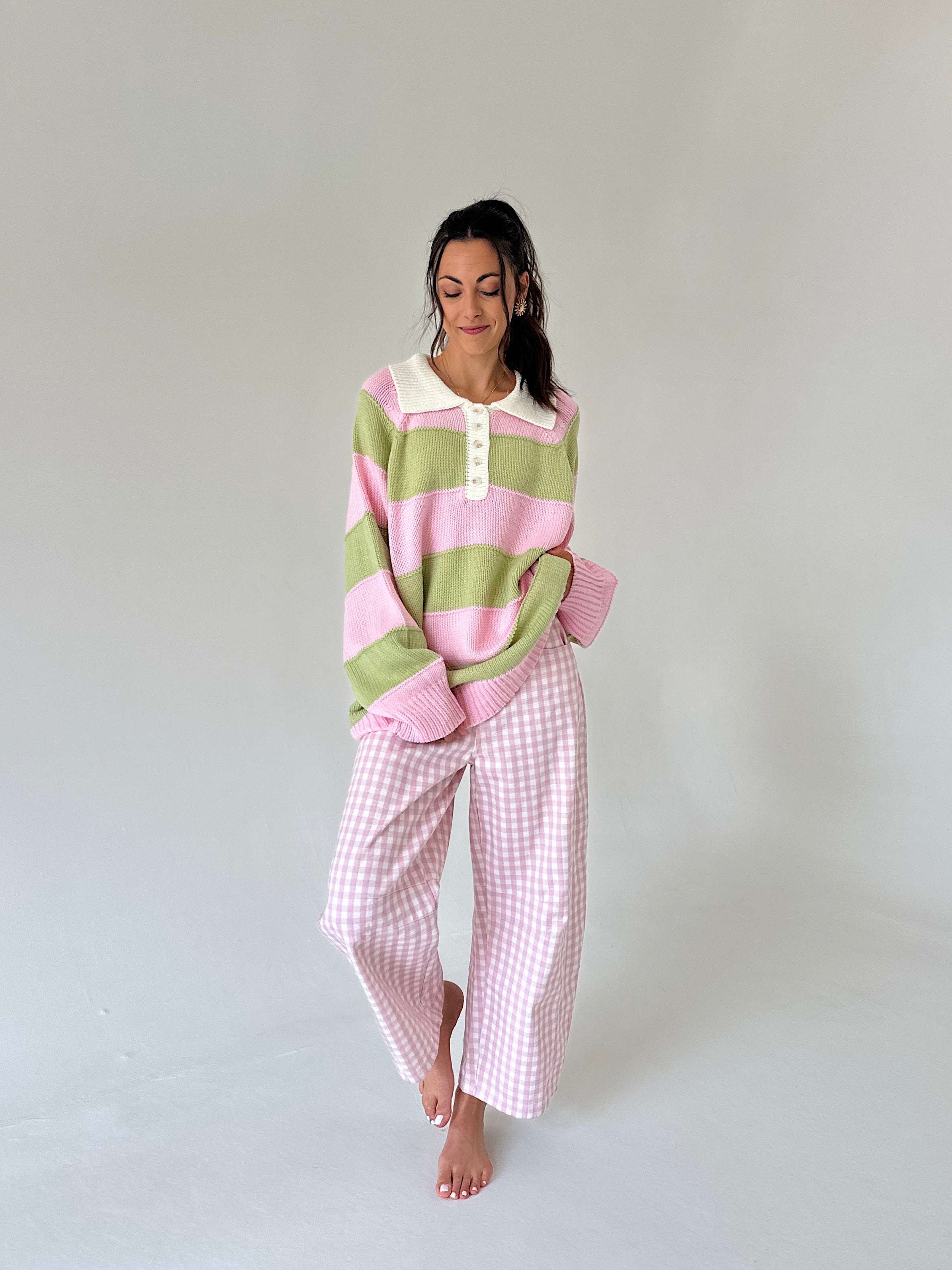 A woman modeling a pink and light green striped henley sweater and matching pink and white gingham wide-leg pants, standing against a neutral light gray studio background.