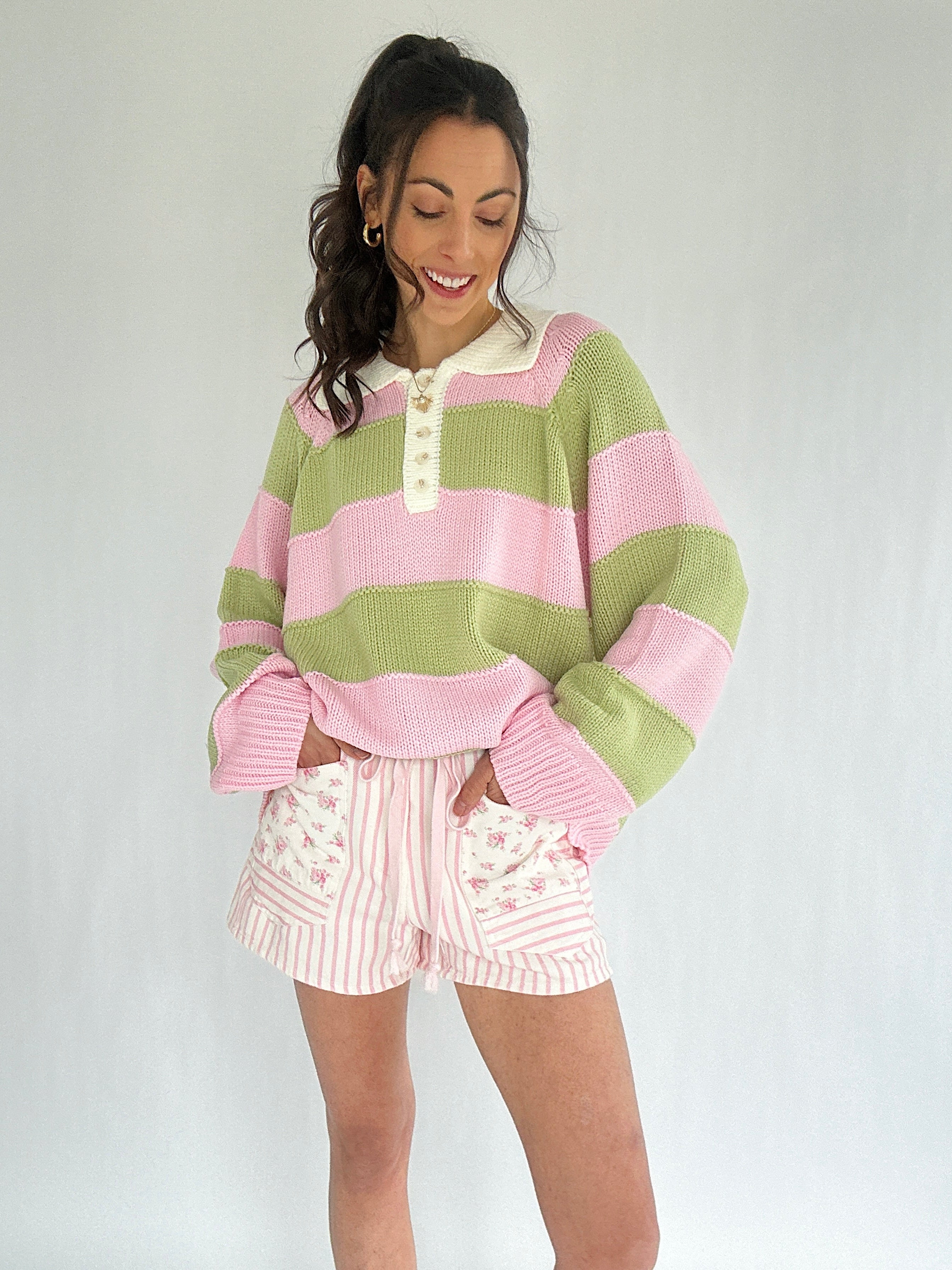 Woman wearing a pink and green striped sweater with white shorts on a plain background