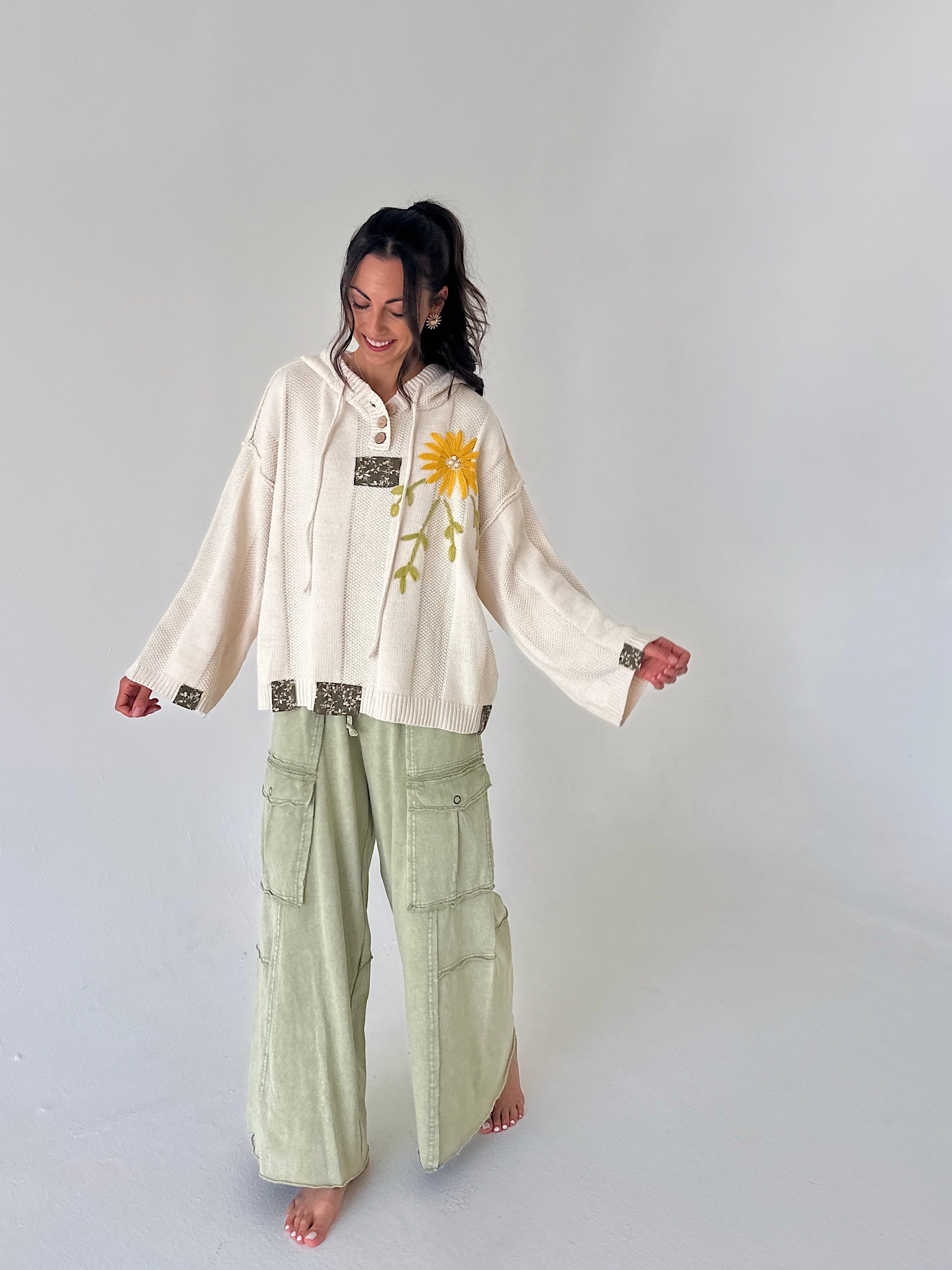 A woman stands barefoot against a light gray studio background, wearing a cream hooded long-sleeve top with a yellow flower graphic and sage green wide-leg cargo pants.