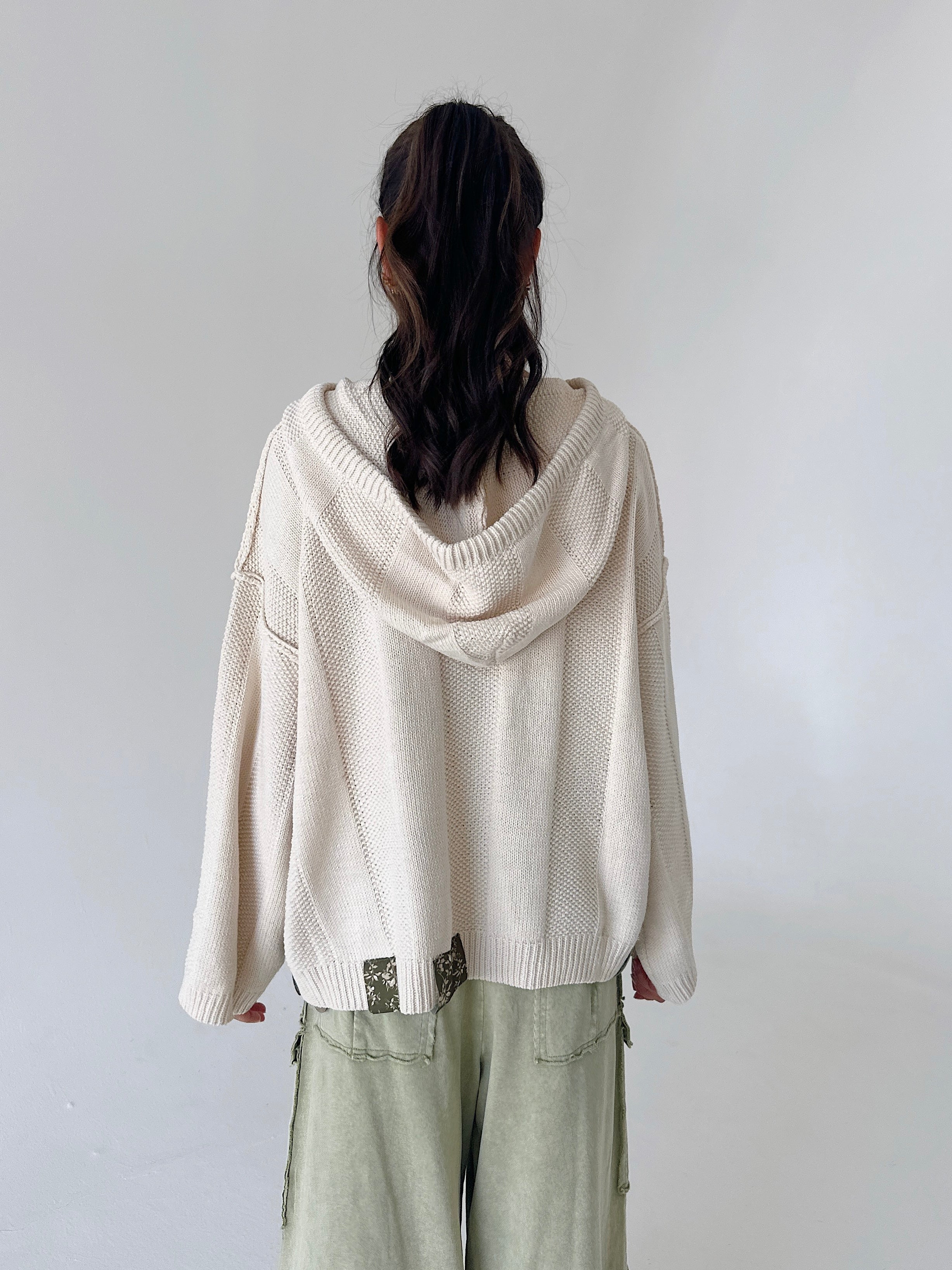 A person shown from the back wearing a cream-colored knit hooded sweater and light sage green pants against a plain light gray background.