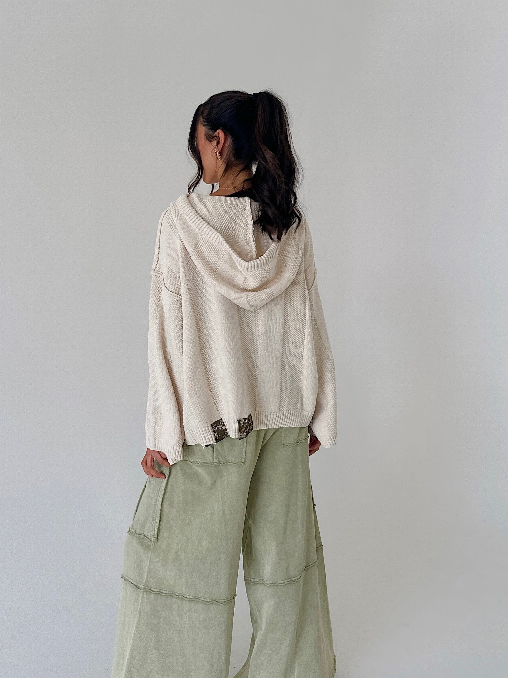 A woman shown from the back wearing a cream-colored knit hooded sweater and light sage green wide-leg pants against a plain light gray background.