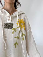 A close-up of a person wearing a cream-colored knit hoodie with a yellow flower and green leaves embroidered on the front, featuring a buttoned collar and a small floral patch pocket.