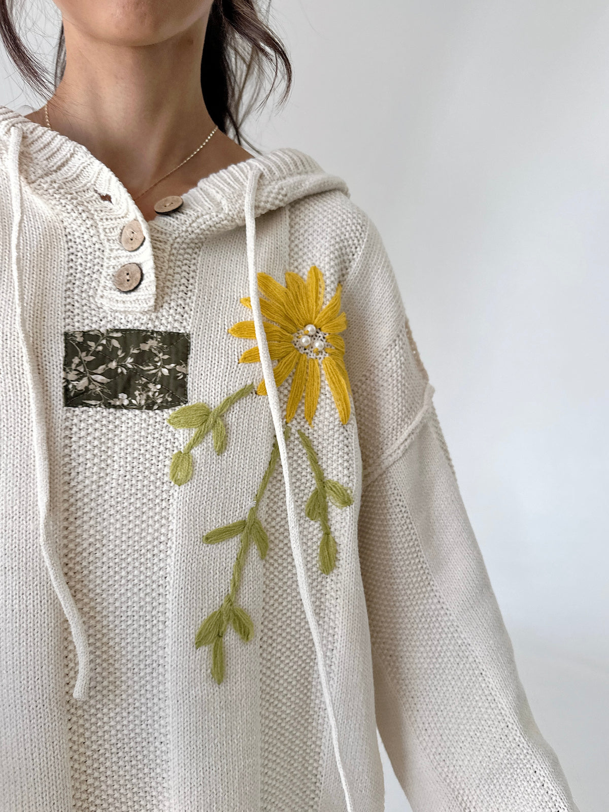 A close-up of a person wearing a cream-colored knit hoodie with a yellow flower and green leaves embroidered on the front, featuring a buttoned collar and a small floral patch pocket.