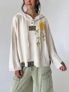 A woman wearing a cream-colored knit hooded sweater with a yellow flower applique on the chest, paired with light sage green cargo pants, against a plain light gray background.