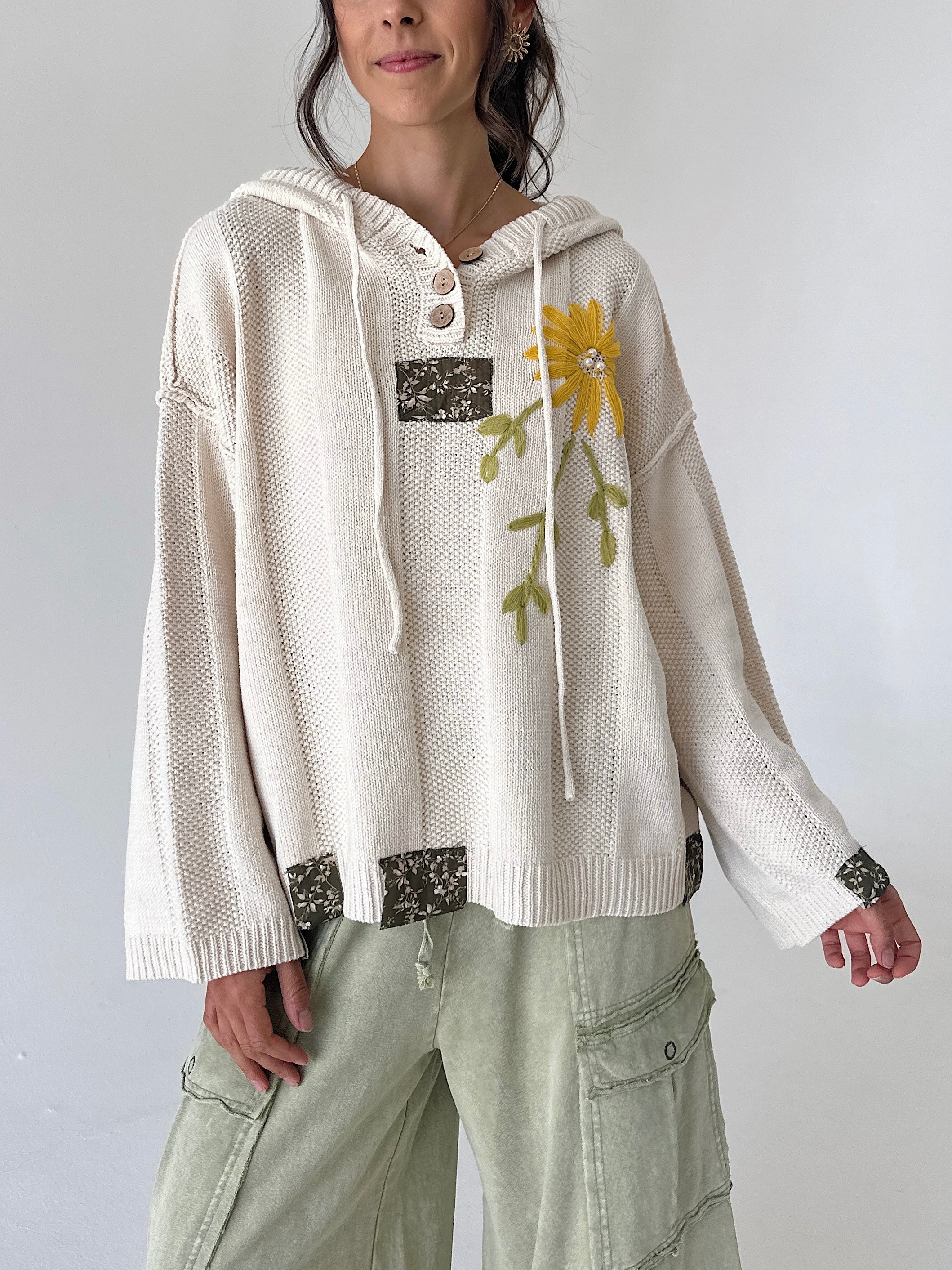 A woman wearing a cream-colored knit hooded sweater with a yellow flower applique on the chest, paired with light sage green cargo pants, against a plain light gray background.