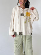 A woman wearing a cream-colored knit hoodie with a yellow sunflower embroidery on the chest, paired with sage green cargo pants, against a plain light gray background.