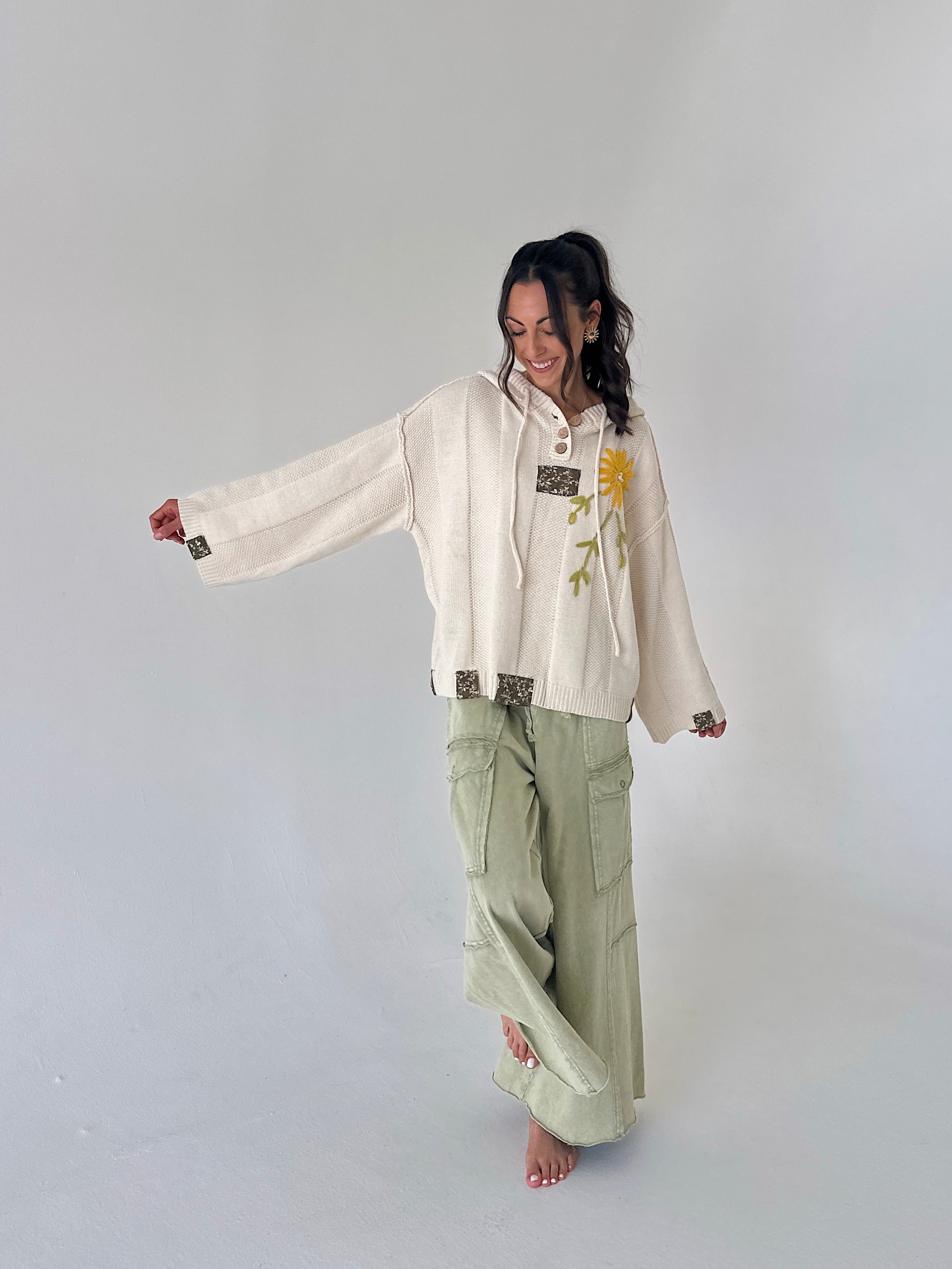 A woman modeling a cream-colored embroidered hoodie with a yellow floral design and sage green wide-leg cargo pants, standing against a plain light gray studio background.