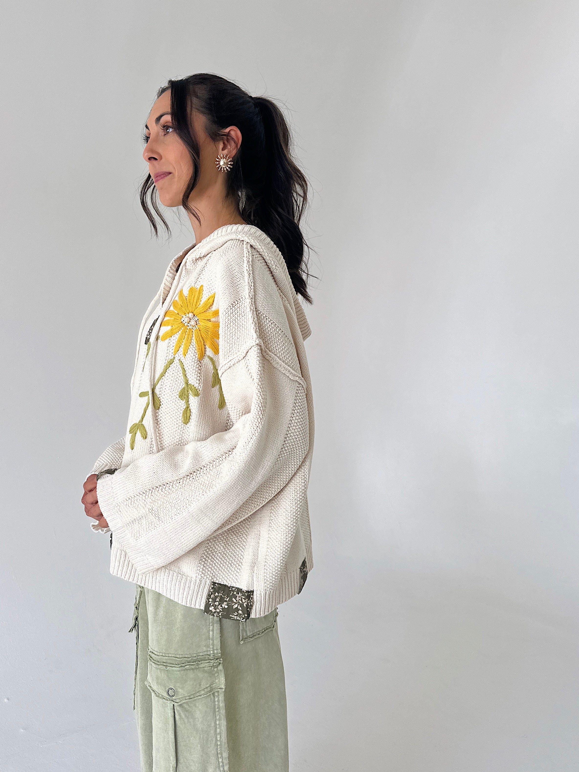 A woman wearing a cream-colored knit hoodie sweater with a yellow sunflower embroidery on the front, paired with sage green cargo pants, standing against a plain light gray wall.