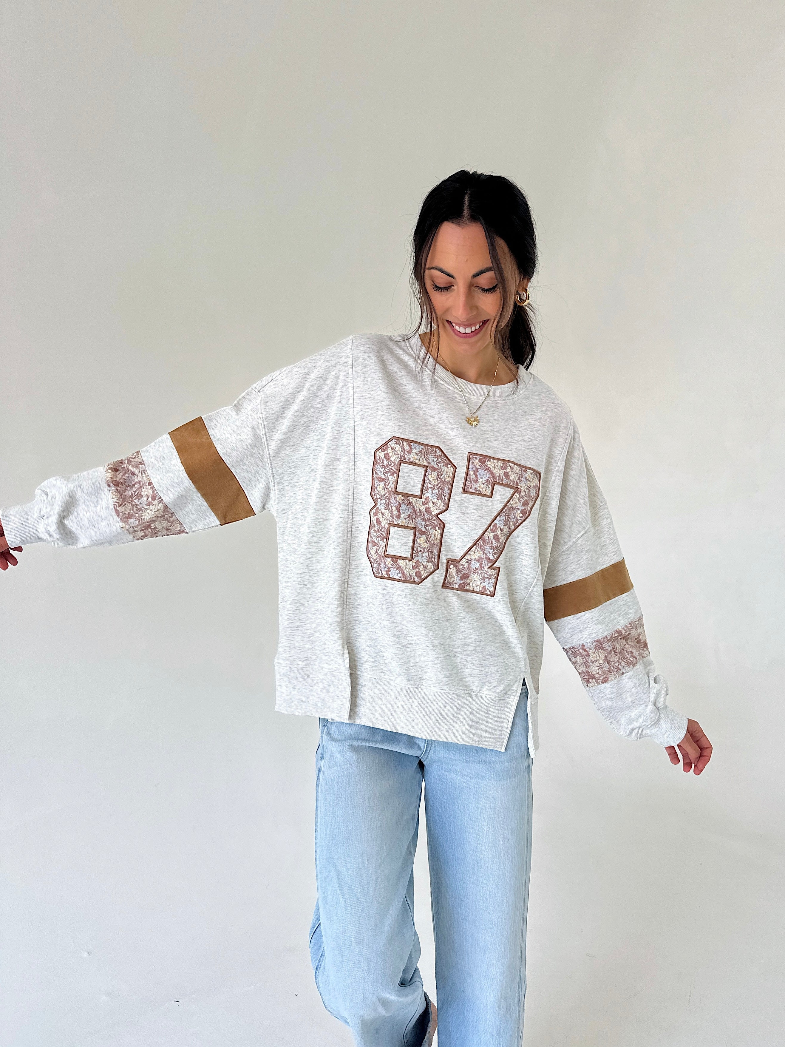 A woman wearing a white heathered long-sleeve sweatshirt with a large '87' graphic on the front, featuring brown and white patterned sleeve stripes, paired with light blue jeans, against a plain off-white background.