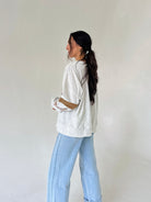 A woman with long dark hair tied back is shown from the side, wearing a heather grey long sleeve top and light blue wide-leg jeans against a plain off-white background.