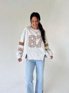 A woman modeling a white heathered long-sleeve sweatshirt with a large '87' graphic on the front, featuring lace-like detailing on the sleeves and light blue jeans, set against a plain off-white studio background.