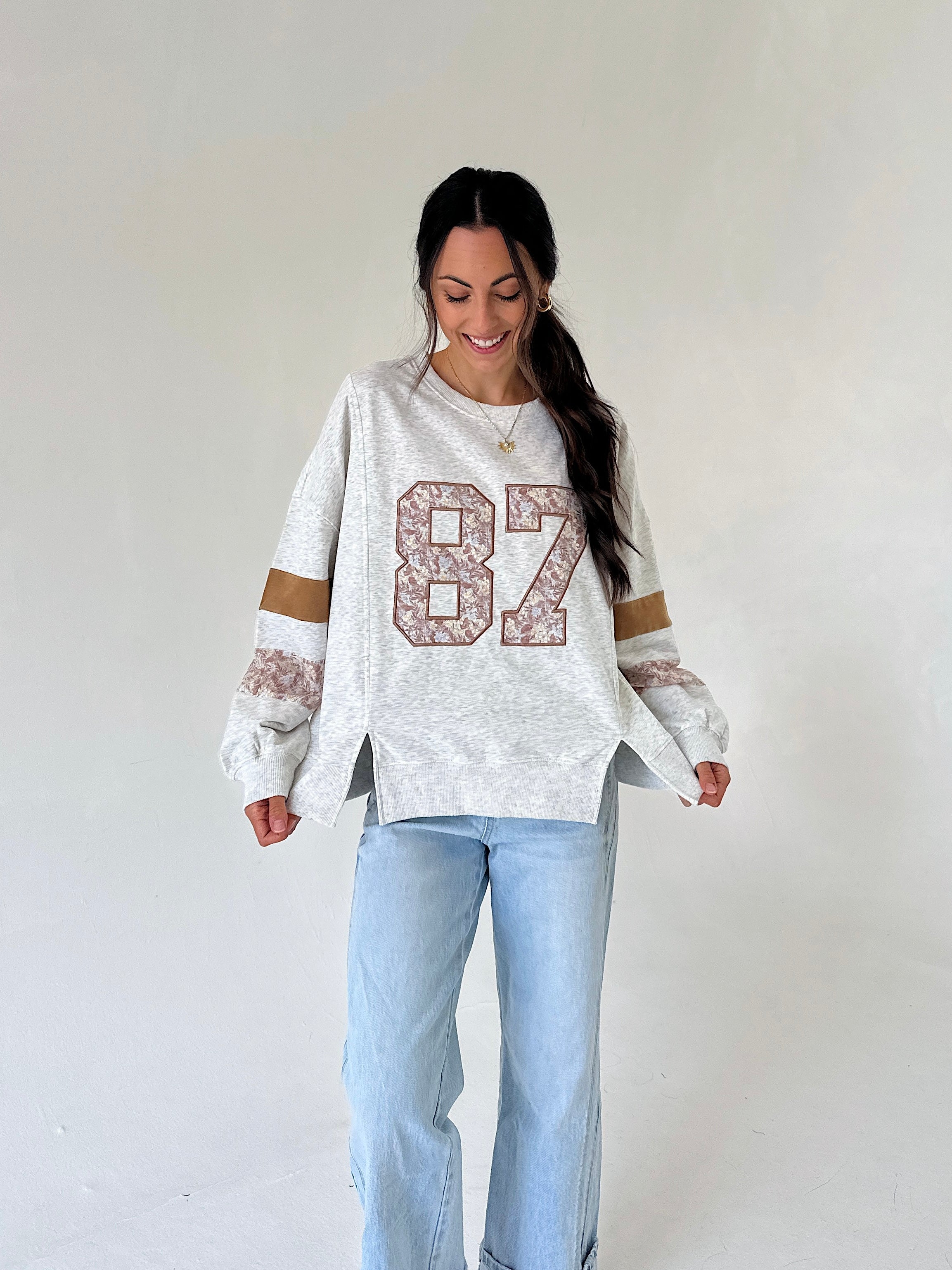 A woman modeling a white heathered long-sleeve sweatshirt with a large '87' graphic on the front, featuring lace-like detailing on the sleeves and light blue jeans, set against a plain off-white studio background.