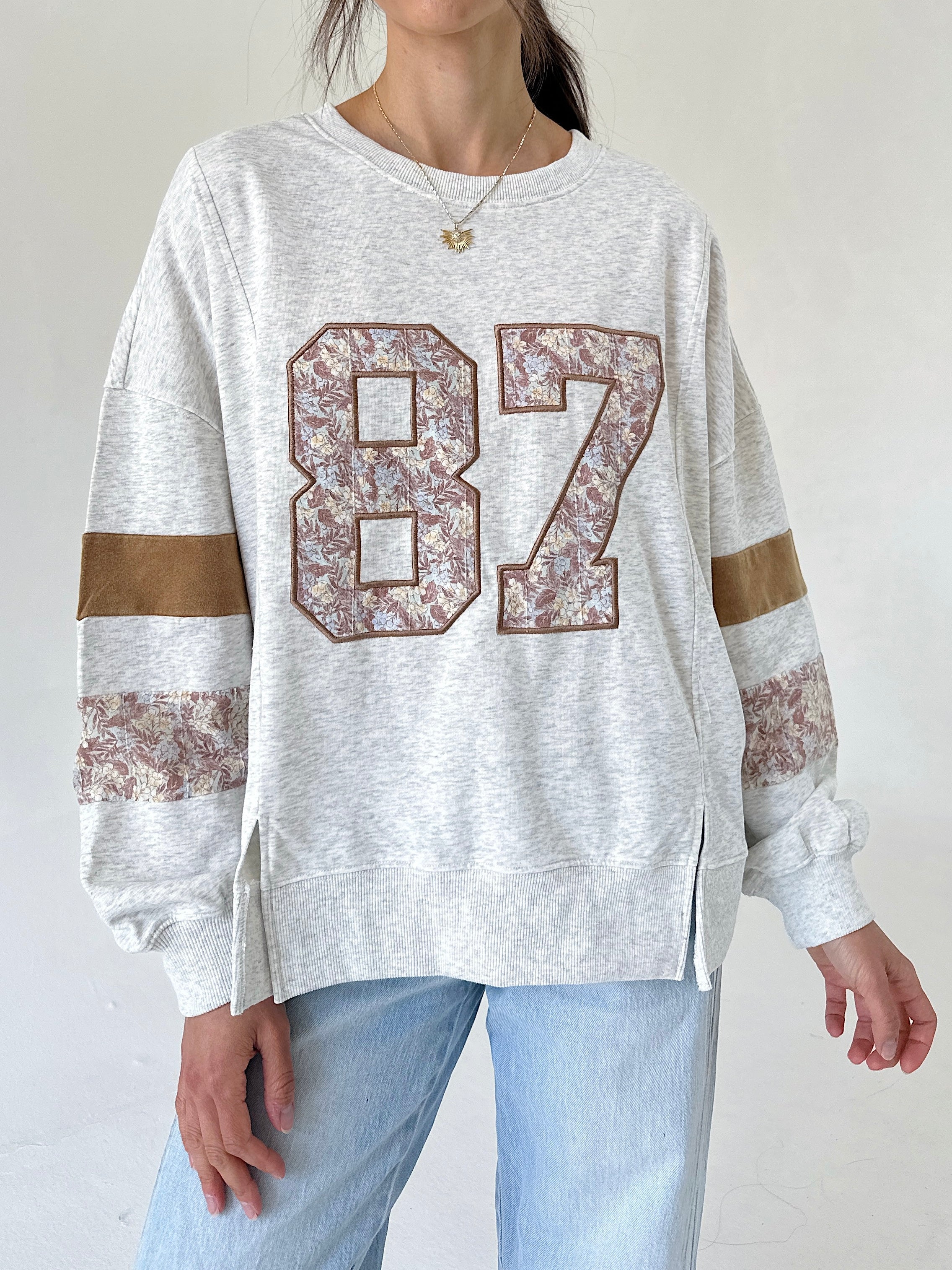 A woman wearing a light heather gray sweatshirt with a large floral-patterned '87' on the front, brown sleeve stripes, and light blue jeans, shown from the neck down against a plain light background.