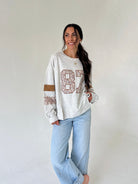 A woman modeling a light heather gray long-sleeve sweatshirt with a distressed brown and beige '87' graphic on the front, paired with light-wash blue jeans, against a plain off-white studio background.